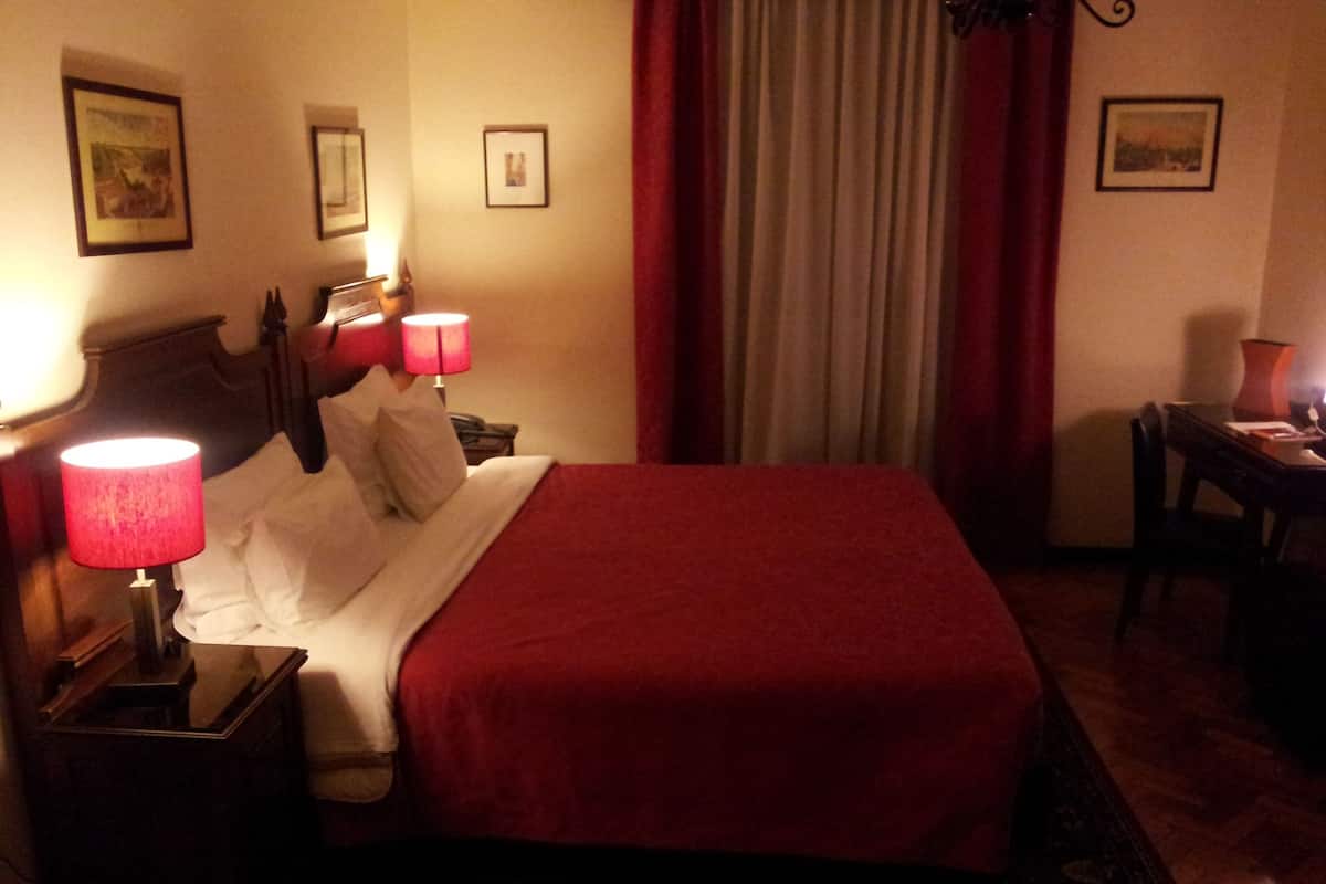 A neatly made bed with a red blanket, two bedside lamps, a wooden nightstand, a small table with a lamp, and a chair.