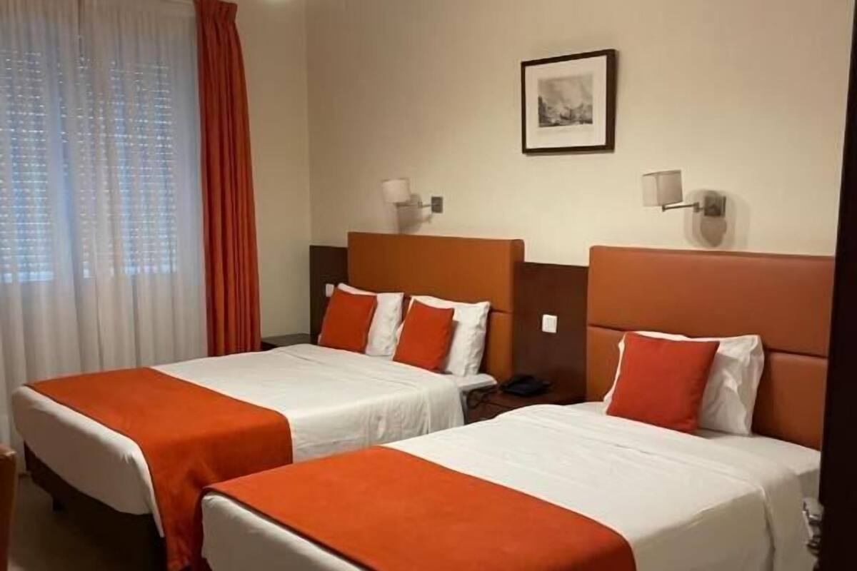 A hotel room with two beds, each with an orange bedspread and white pillows. There is a ceiling light fixture above the beds, a framed picture on the wall, and a window with white curtains.