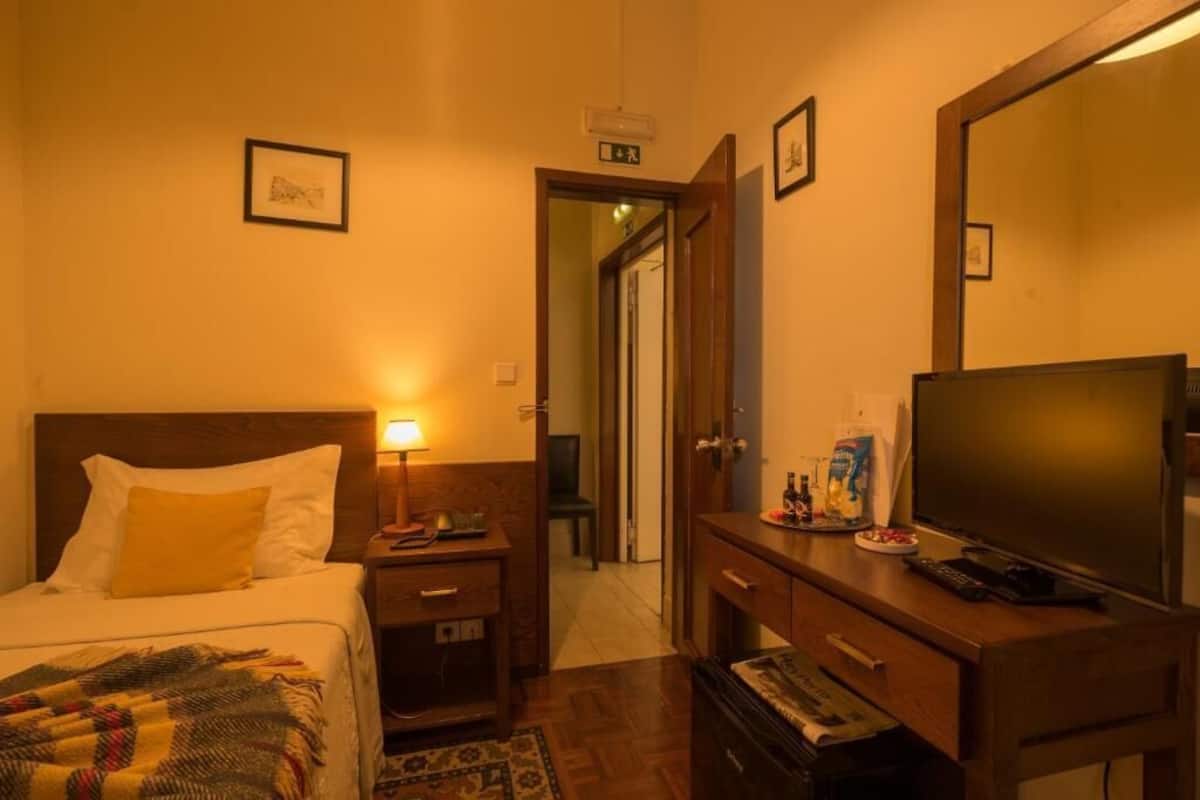 A hotel room with a bed, bedside table, lamp, TV, and a desk with a computer.