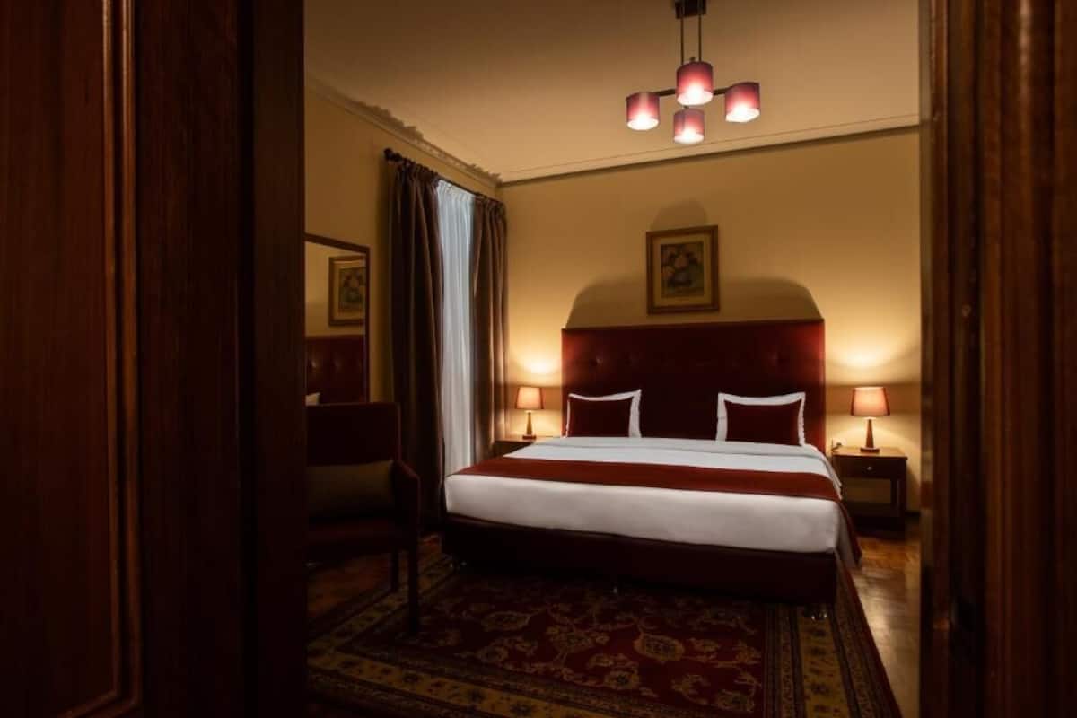 A hotel room with a large bed, two bedside tables with lamps, a red headboard, a patterned rug, and a chandelier.