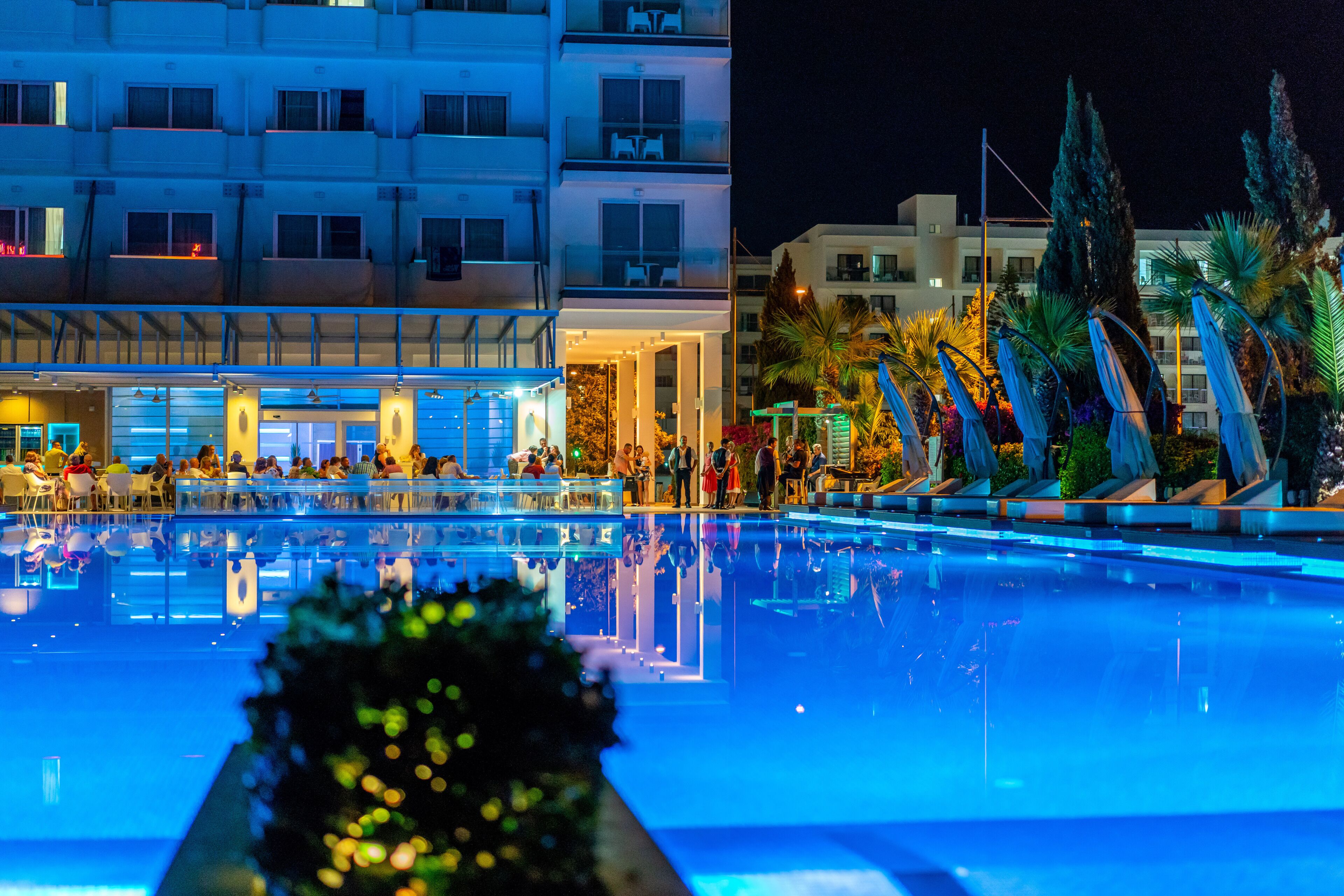 A nighttime view of a hotel pool area with a clear pool, outdoor seating, and a building with multiple balconies.