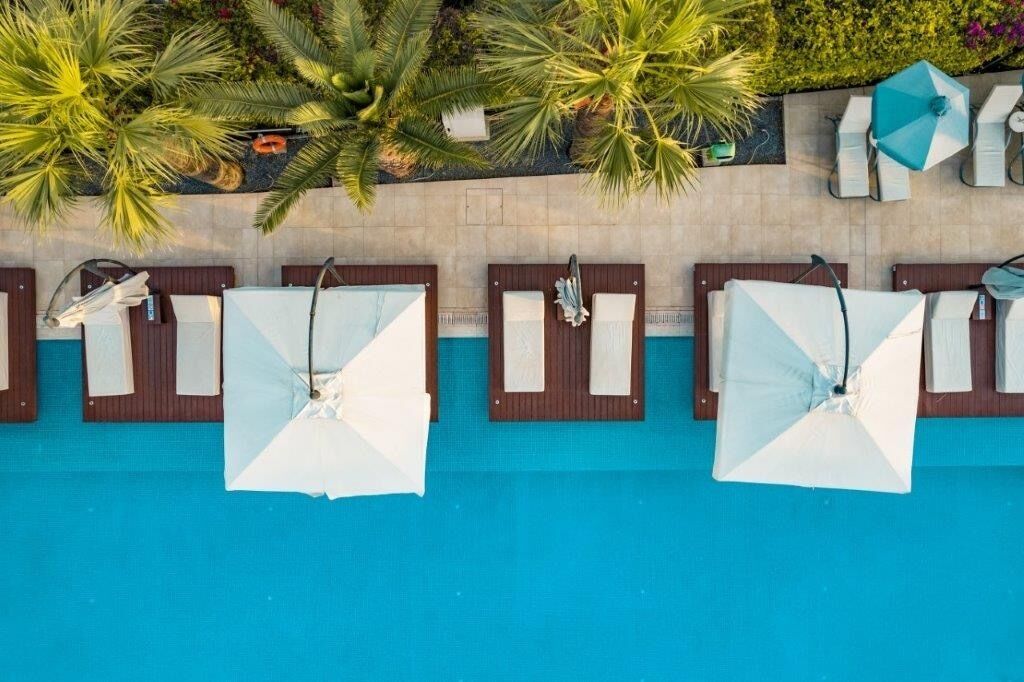 Vista aérea de una zona de piscina con numerosas tumbonas y sombrillas, rodeada de palmeras y un paseo de baldosas.