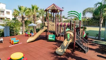 A playground with slides and climbing structures, surrounded by palm trees and a tennis court.