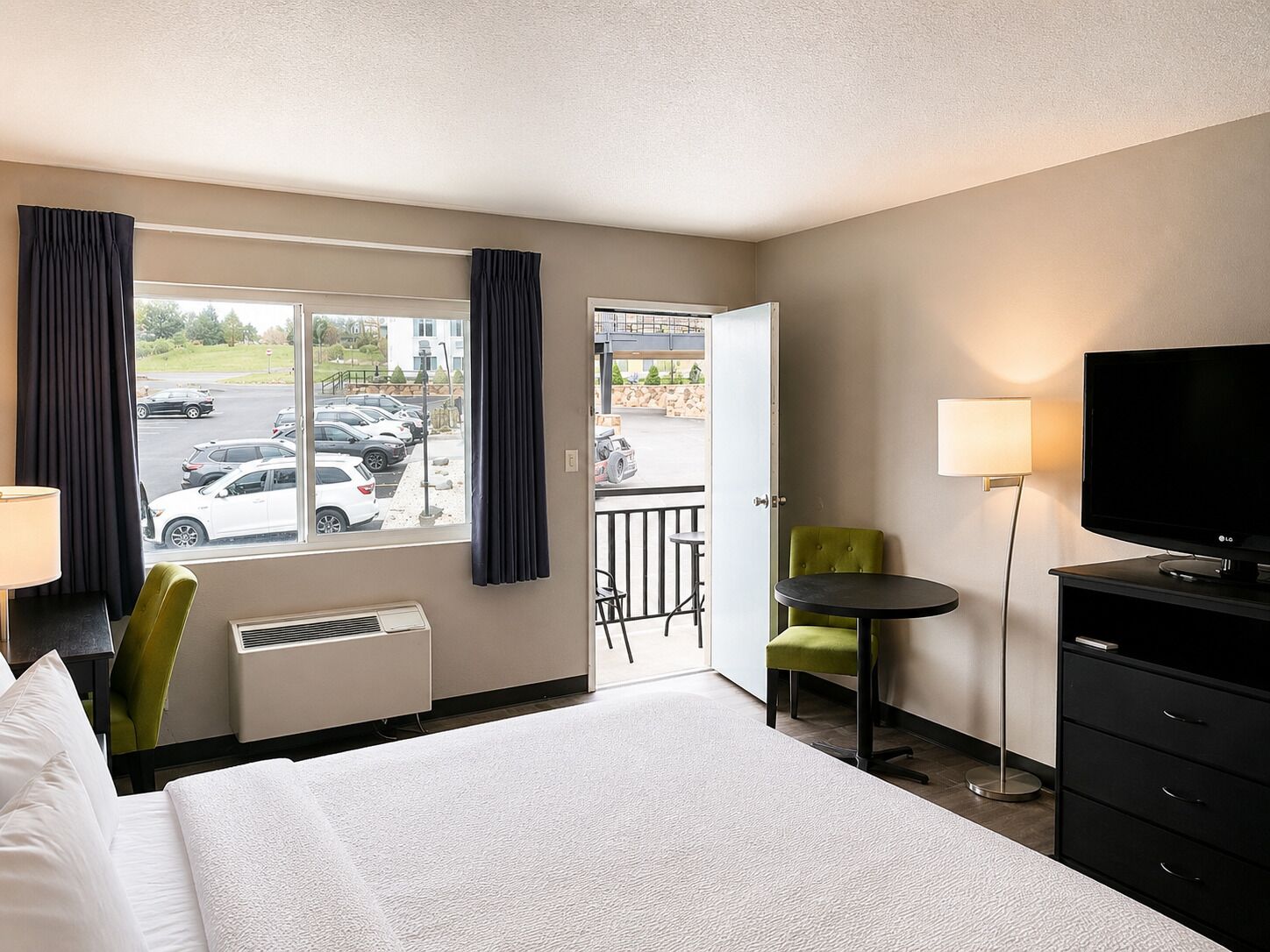 A hotel room with a bed, a desk, a chair, a TV, and a view of parked cars through the window.