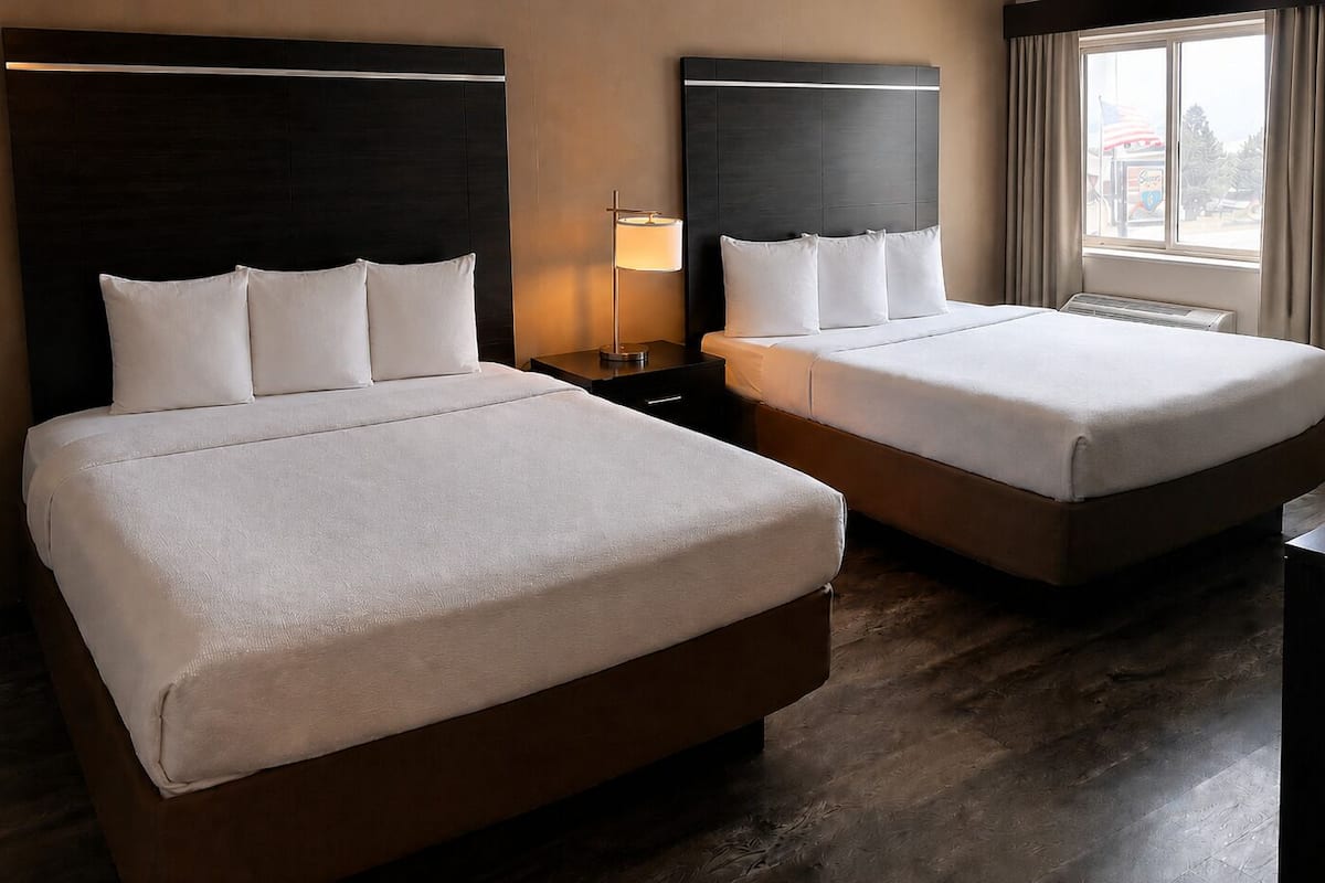 A hotel room with two beds, a nightstand with a lamp, a window with curtains, and a view of the outside.
