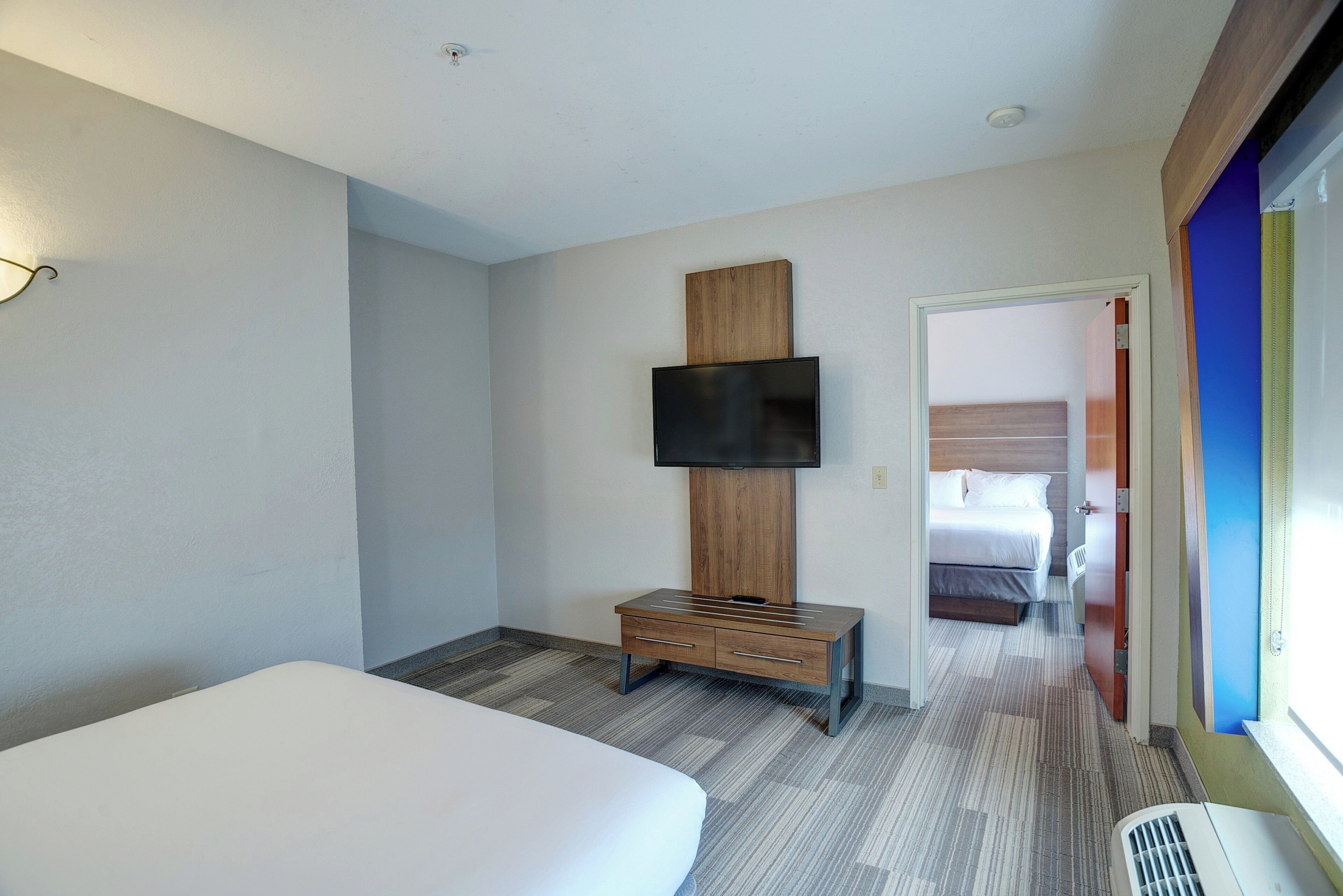 A hotel room with a bed, a TV mounted on a wooden panel, a small wooden bench, and a door leading to another room.