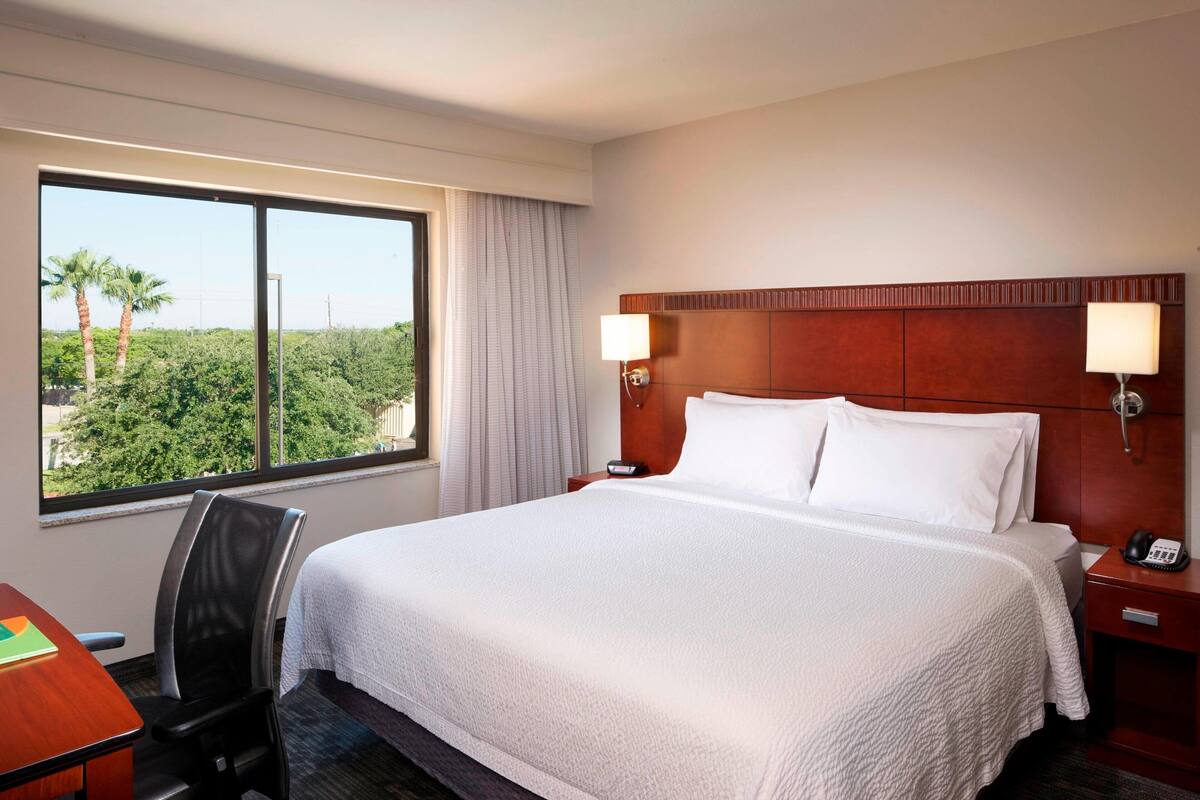 A hotel room with a large bed, two bedside lamps, a desk with a chair, and a window with a view of greenery.