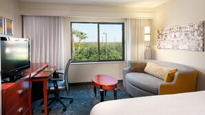 Habitación de hotel con cama, escritorio con televisor, silla, mesita pequeña y ventana grande con vista a follaje.