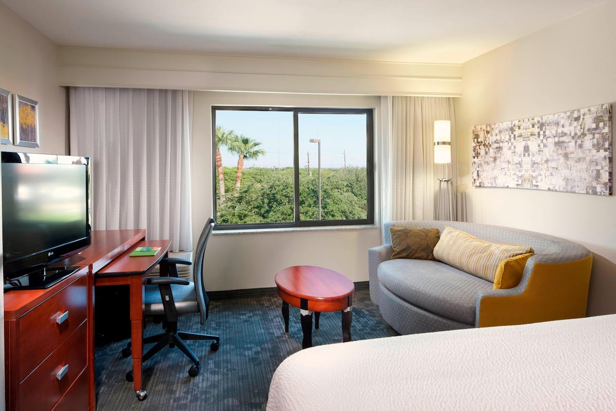 A hotel room with a bed, a desk with a TV, a chair, a small table, and a large window with a view of greenery.