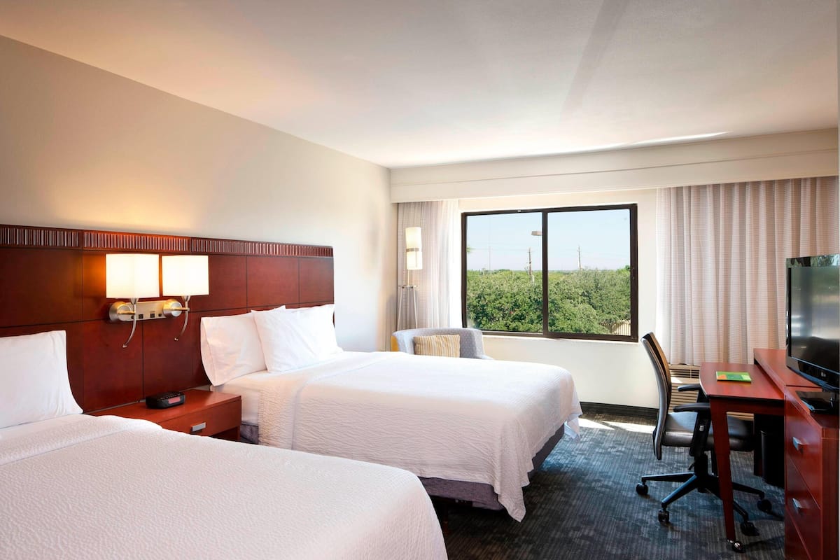 A hotel room with two beds, a desk with a chair, a TV, and a large window with a view of greenery.