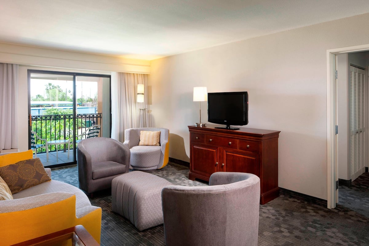 Une chambre d’hôtel équipée d’un canapé, d’un fauteuil, d’un pouf, d’un téléviseur posé sur un meuble en bois et d’un balcon donnant sur un paysage verdoyant.