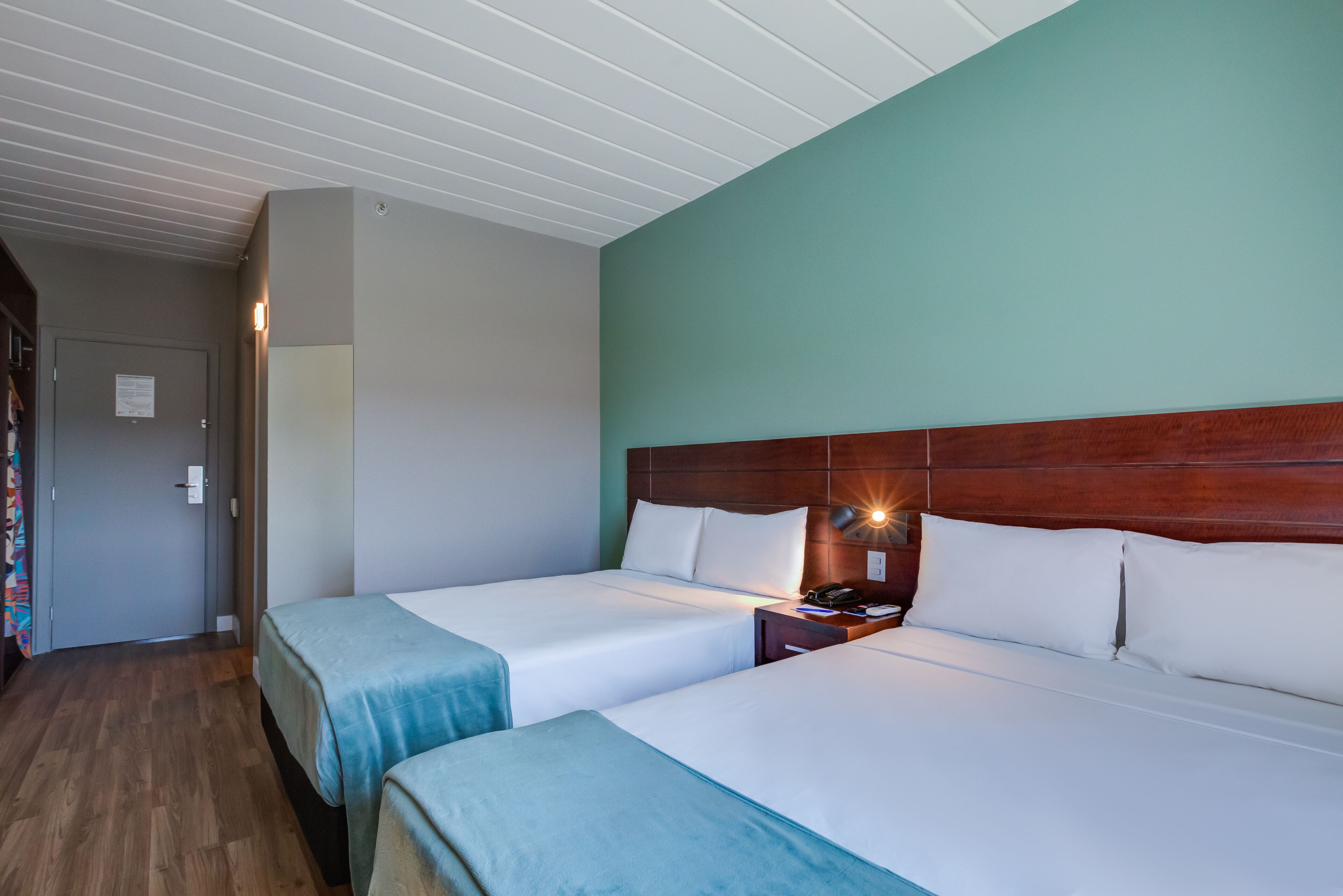 Habitación de hotel con dos camas individuales, cabeceros de madera, una mesita de noche con lámpara y una puerta con un cartel.