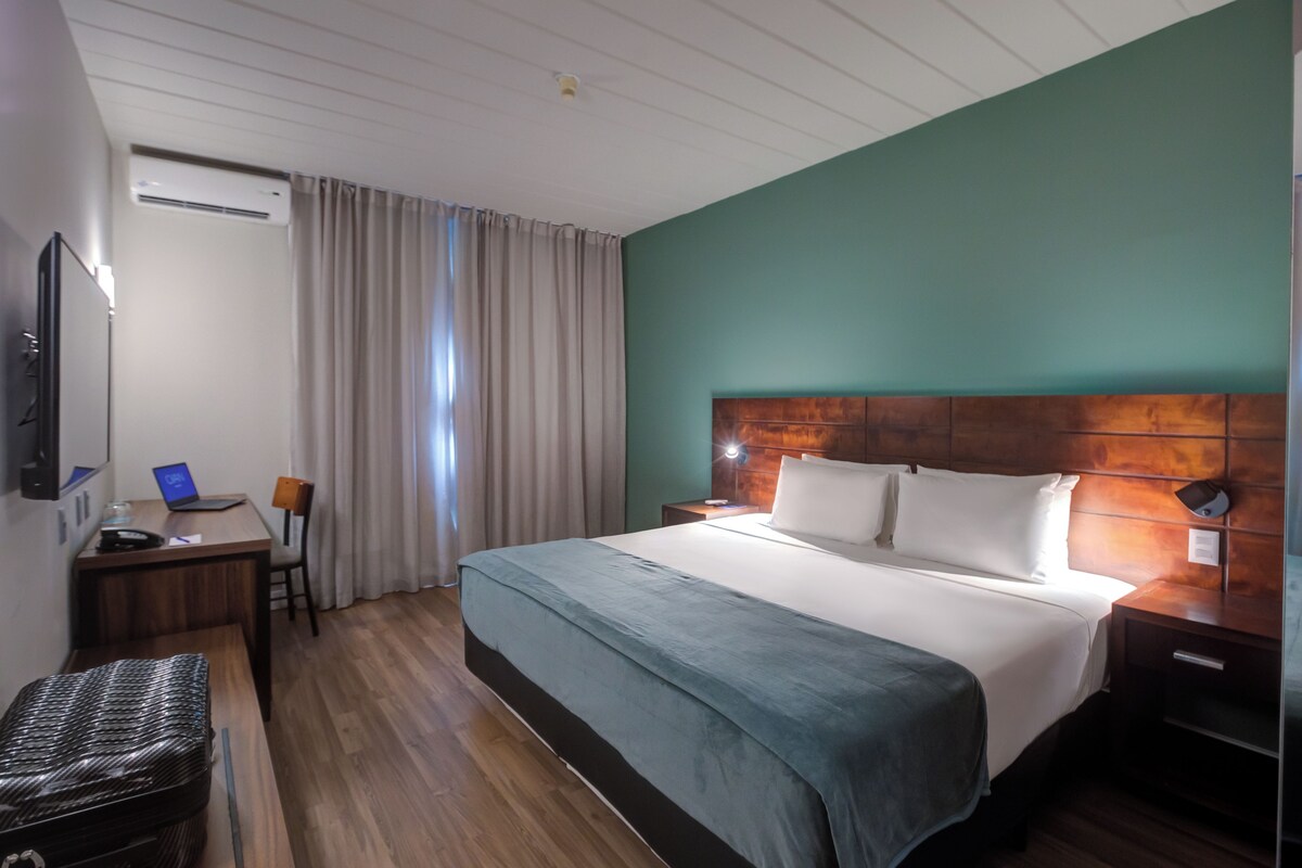 A hotel room with a bed, desk, chair, and a suitcase.