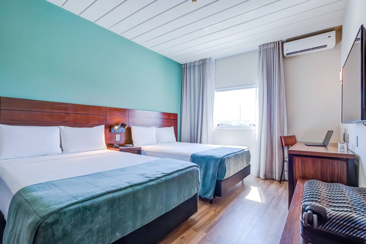 Ein Hotelzimmer mit zwei Betten, einem Schreibtisch mit einem Laptop, einer Klimaanlage und einem Fenster mit Vorhängen.