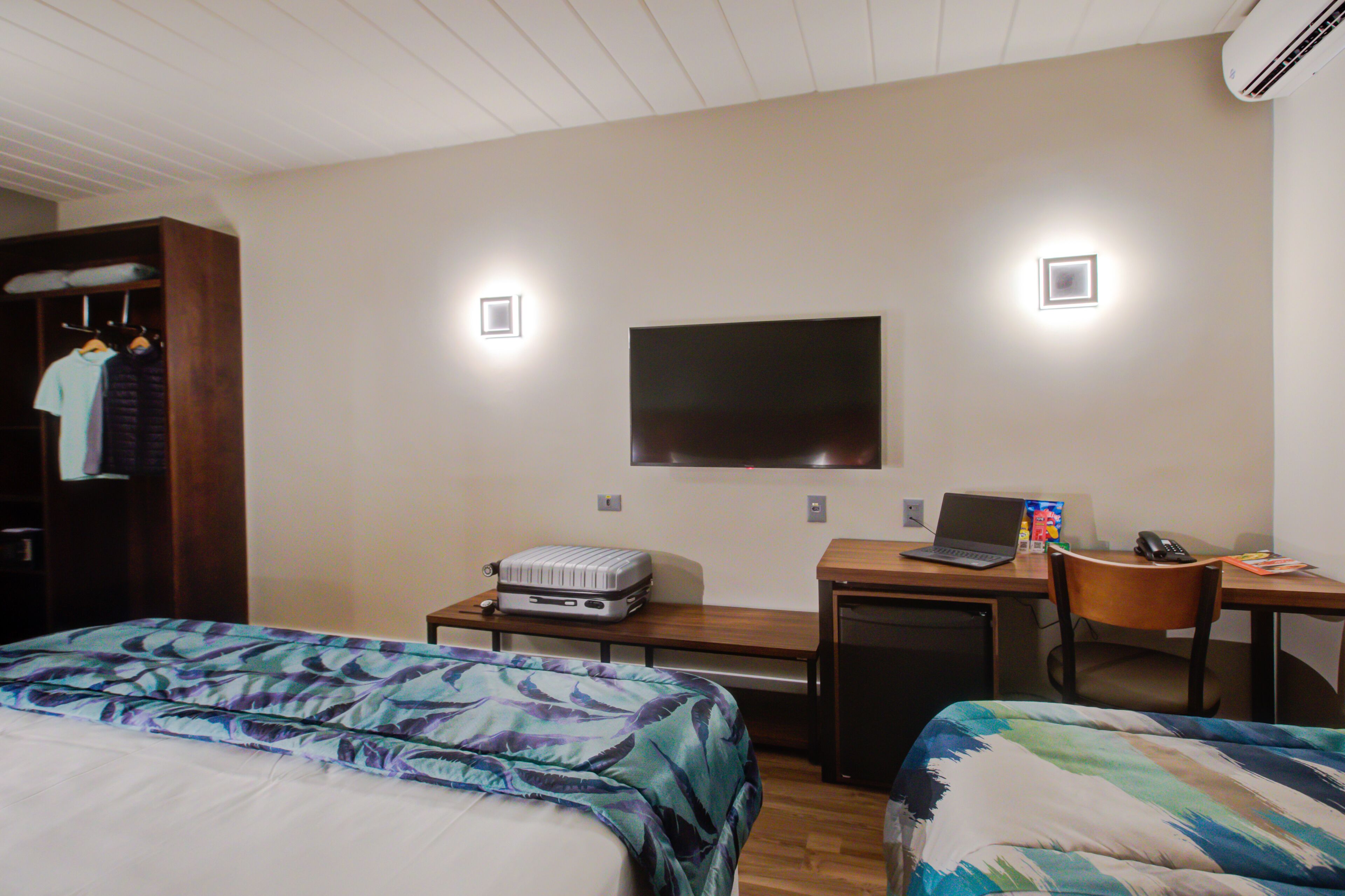 Ein Hotelzimmer mit zwei Betten, einem Schreibtisch mit einem Laptop, einem Koffer und einem Fernseher an der Wand.