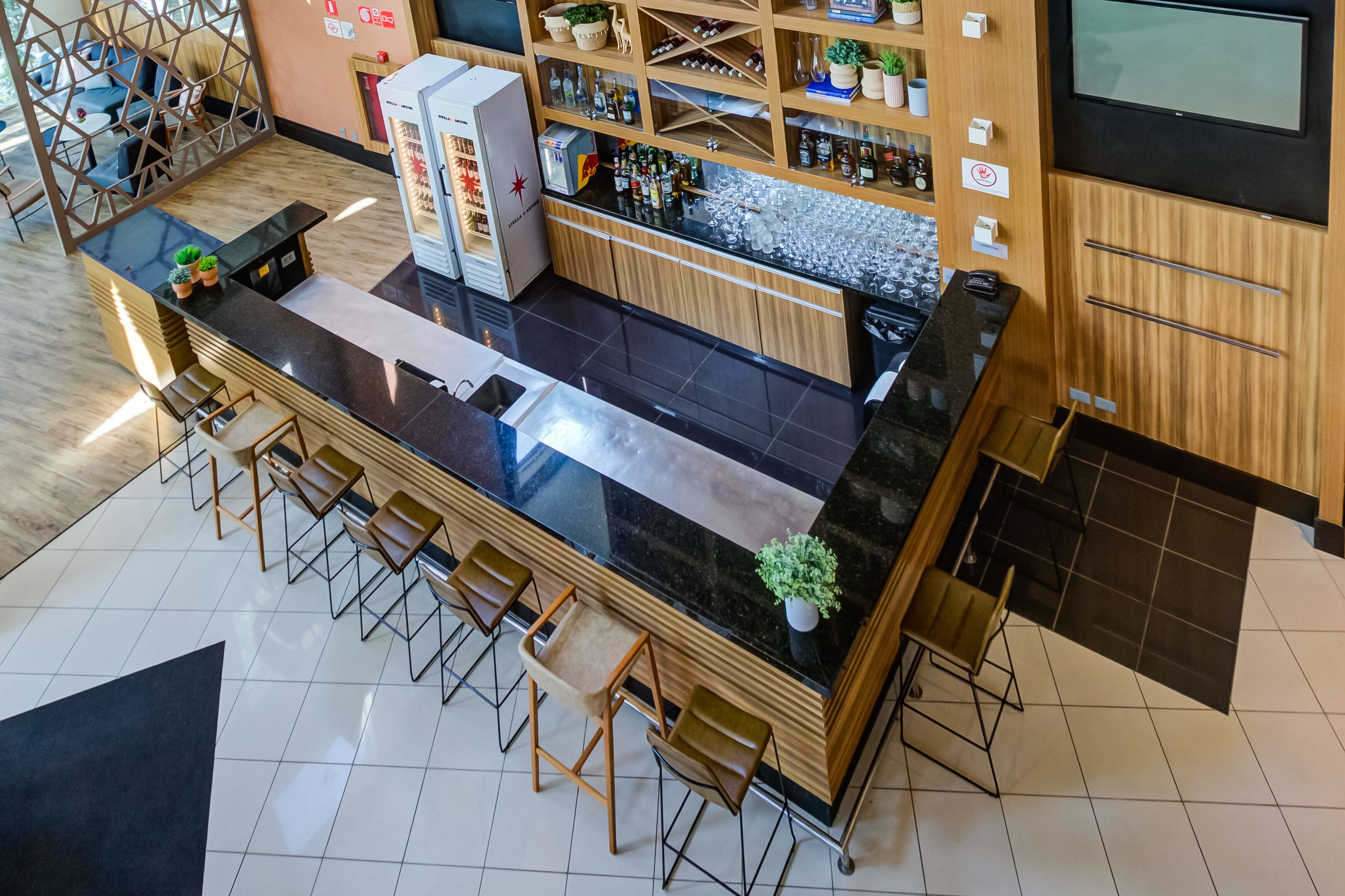 Zona de bar moderna con mostrador negro, taburetes de madera y una variedad de botellas y vasos.