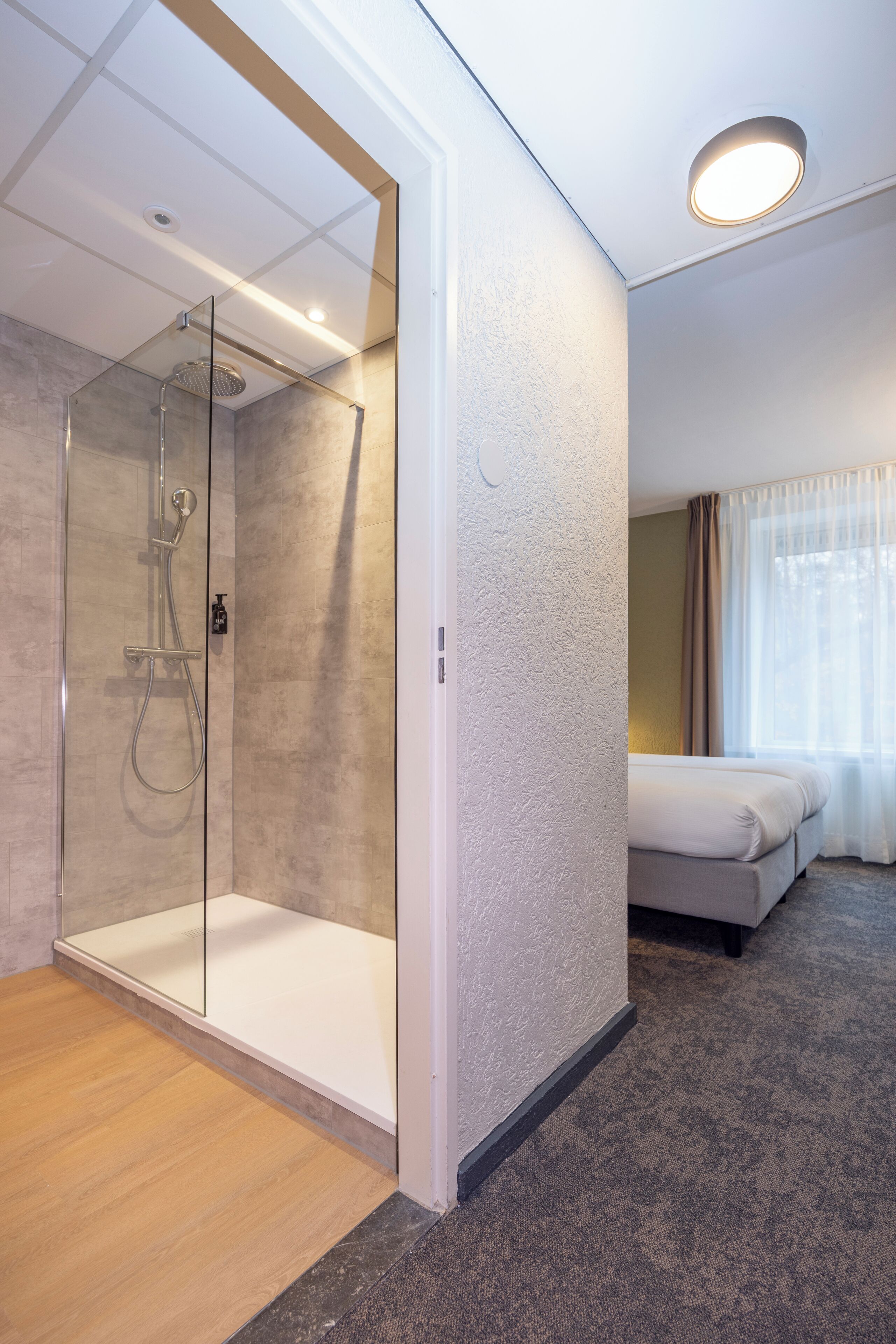 A modern hotel room with a glass-enclosed shower, a bed, and a window with sheer curtains.