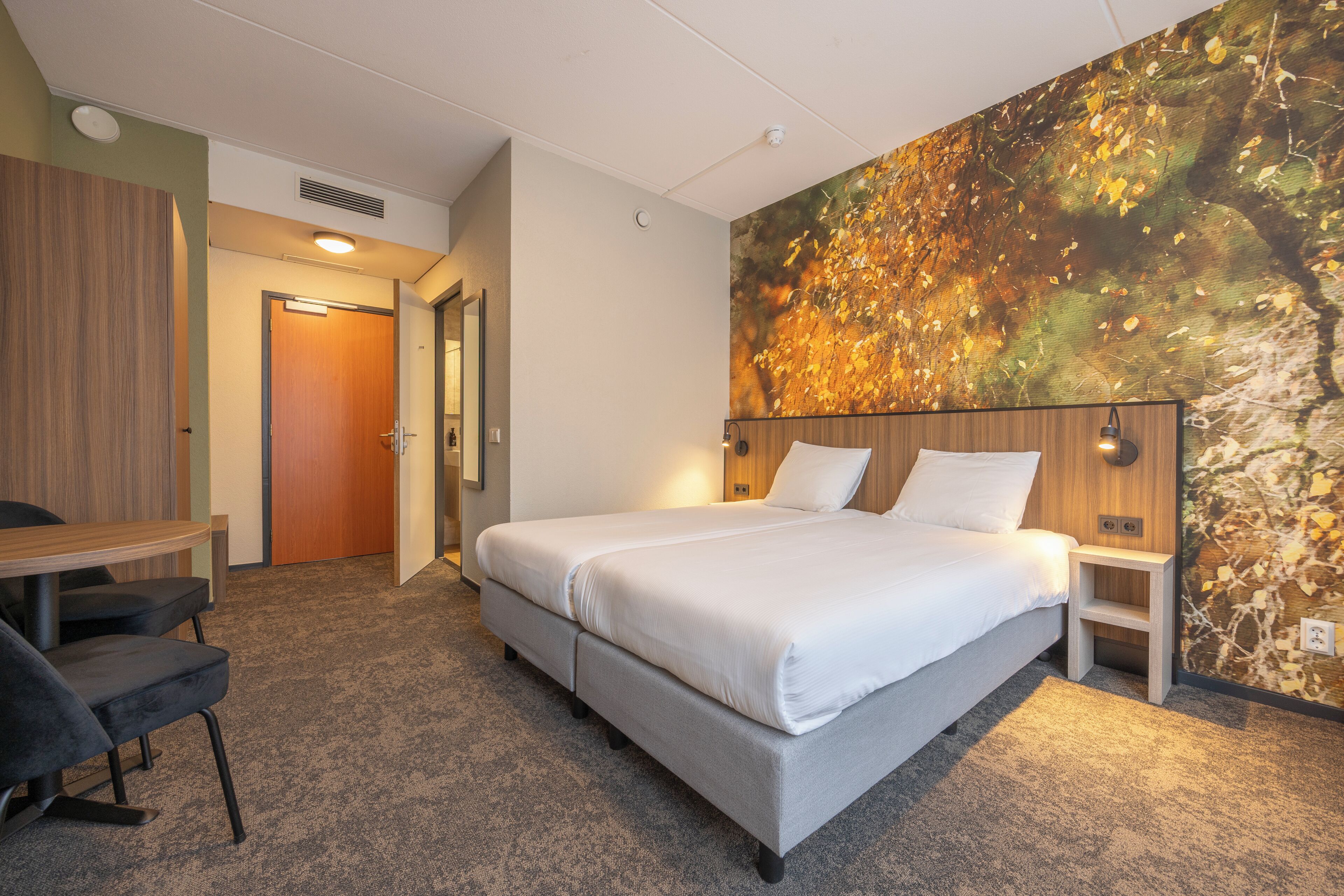 A hotel room with a bed, a desk, and a chair. There is a door leading to another room.