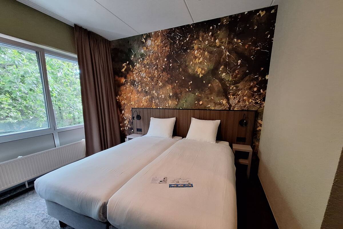 Un lit double avec du linge de lit blanc, une tête de lit ornée d’un motif de feuilles, une table de chevet et une fenêtre avec des rideaux.