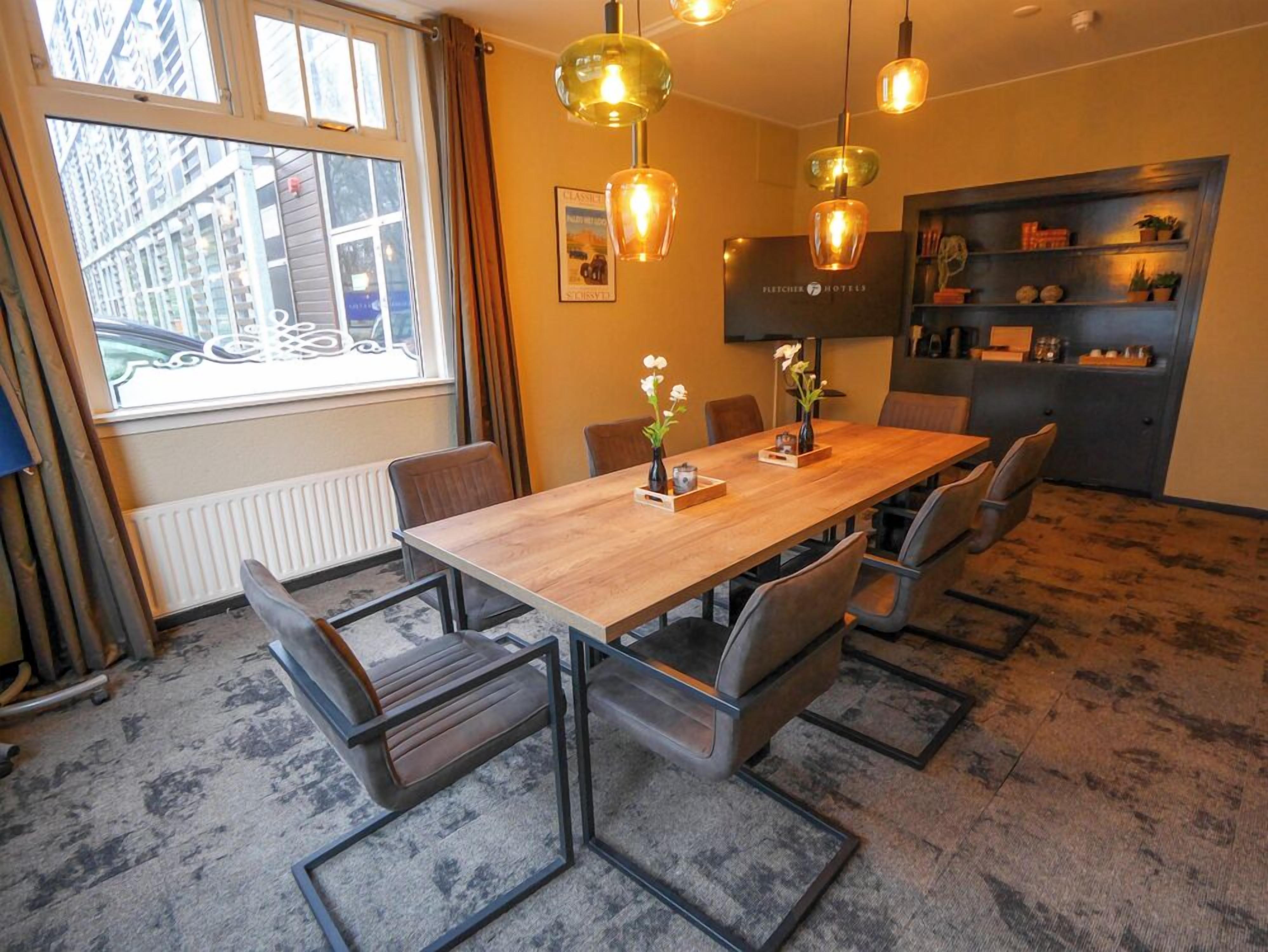 Habitación de hotel con una mesa de comedor de madera, sillas modernas, luces colgantes, un radiador, una ventana con cortinas y una estantería con objetos decorativos.