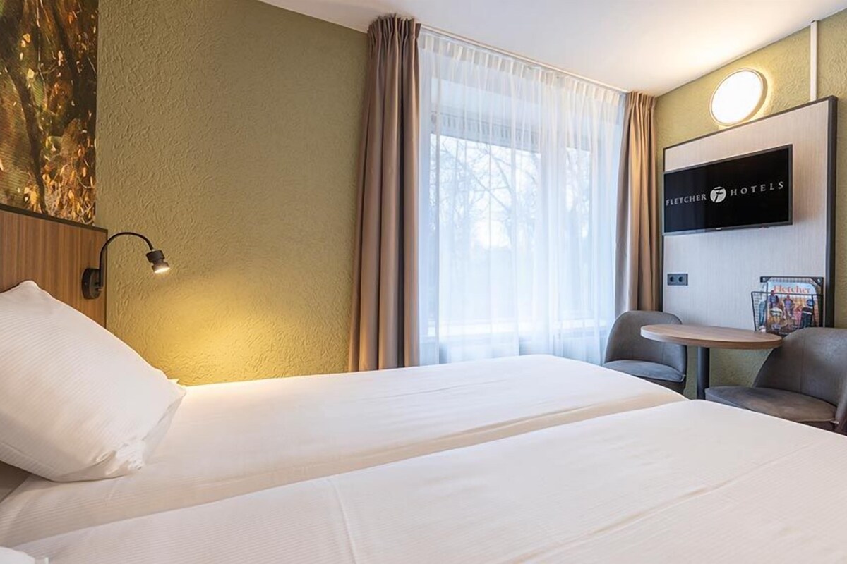 Hotelkamer met een groot bed, een bureau met stoel, een flatscreen televisie en een raam met dunne gordijnen.