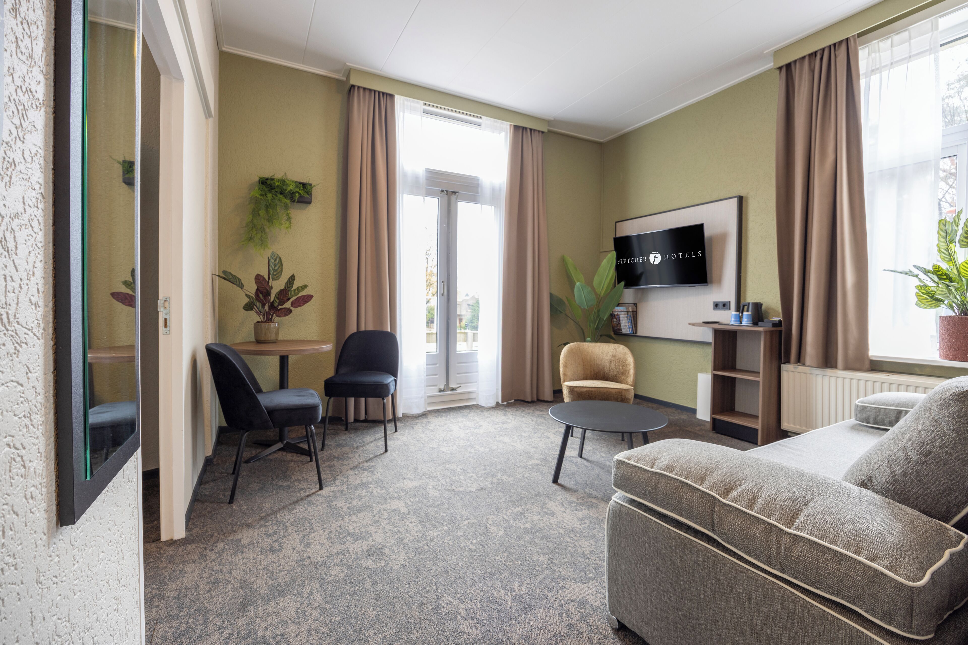 Une chambre d’hôtel moderne avec un canapé, deux fauteuils, une petite table, un téléviseur et une plante en pot.