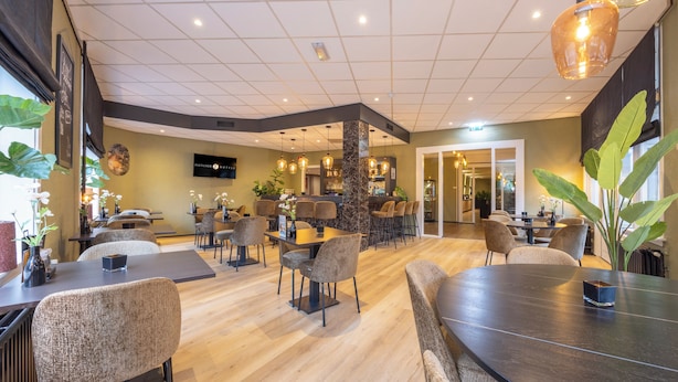 A spacious restaurant with multiple seating areas, including round tables and high chairs. There is a bar area with a counter and stools. The space features a mix of wooden and upholstered furniture, and the ceiling has a grid pattern with recessed lighting. - Fletcher Hotel-Restaurant Apeldoorn (Apeldoorn)