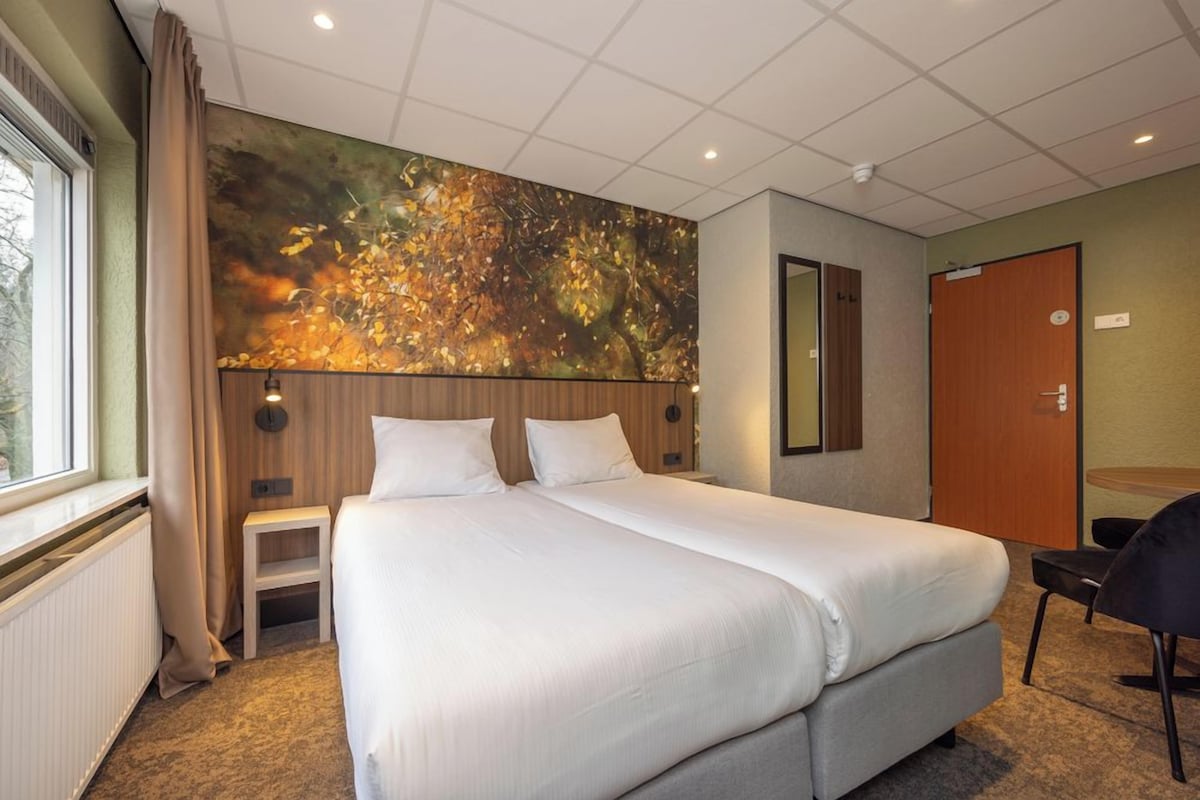 Hotelkamer met een groot bed, een klein nachtkastje, een stoel, een spiegel en een raam met gordijnen.