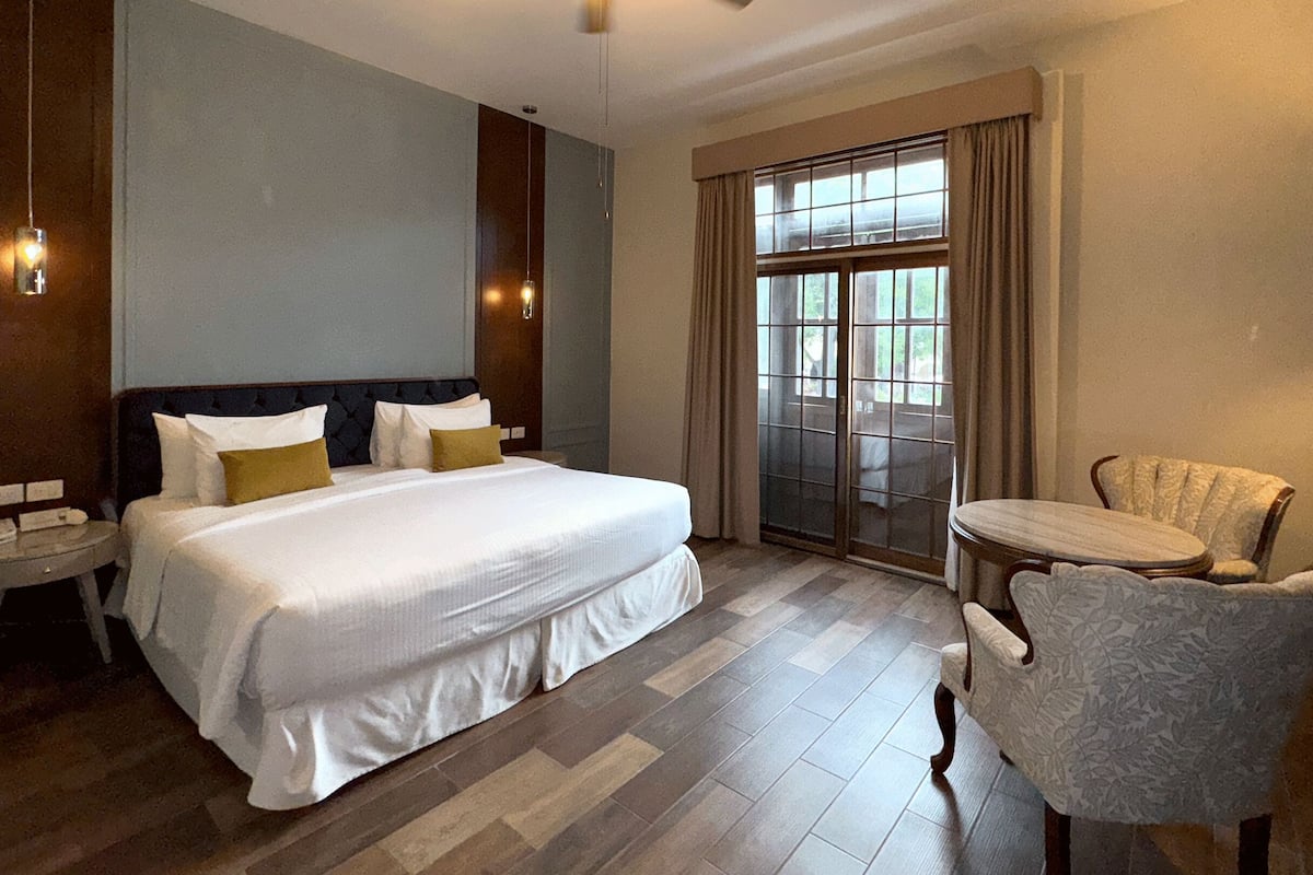 A hotel room with a large bed, a chair, a small table, and a window with curtains.