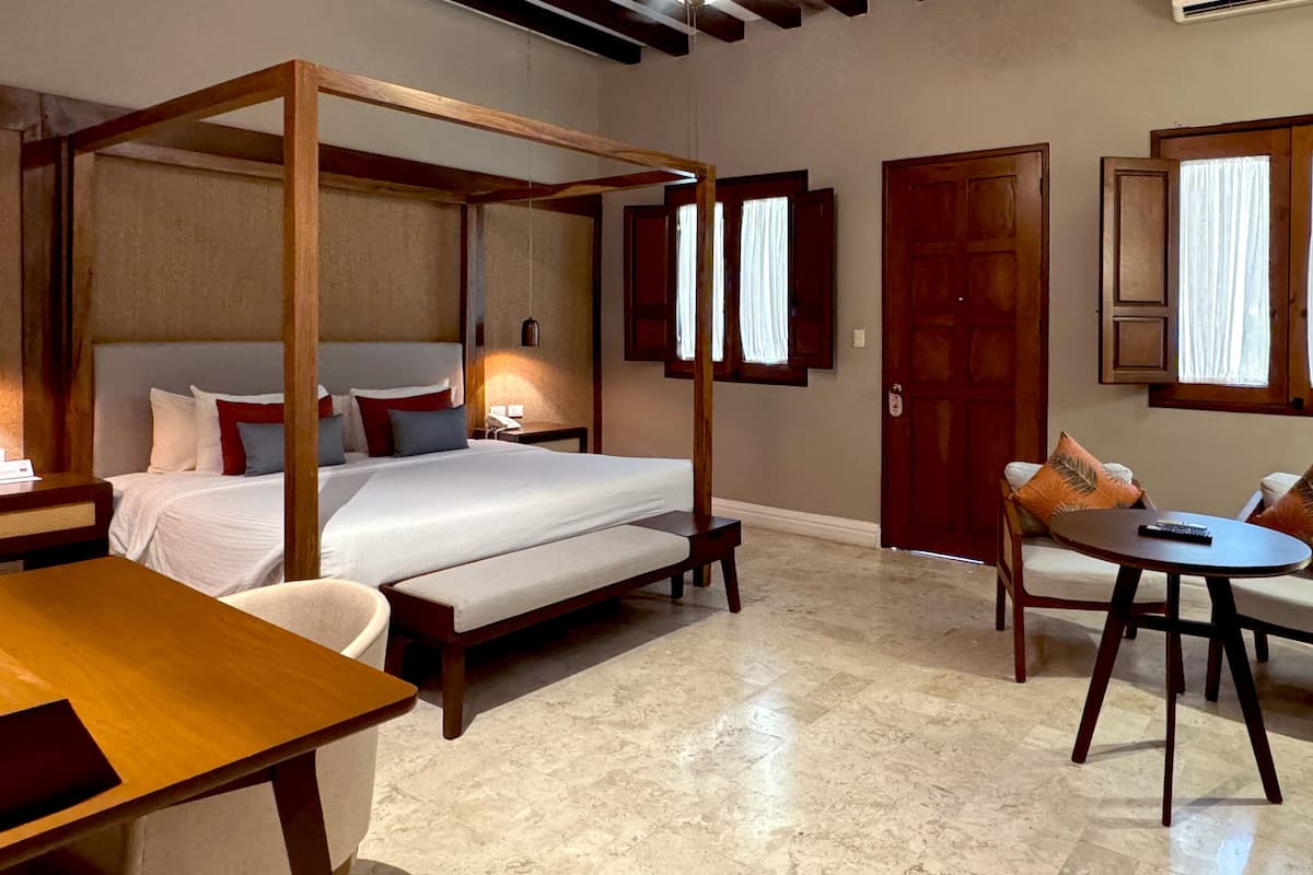 A four-poster bed with a wooden headboard, a wooden desk, a round table, and a sofa with cushions.