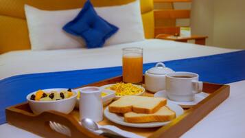 Bandeja de desayuno con tostadas, huevos revueltos, fruta, café y jugo sobre una cama con almohada azul y cabecera de madera.