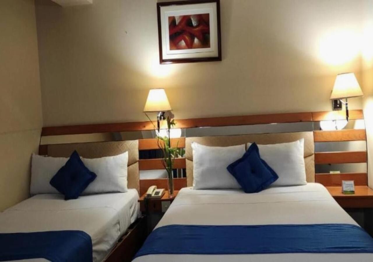 A hotel room with two single beds, each with a blue pillow and white bedding. There are bedside lamps, a small table with a phone, and a framed picture on the wall.