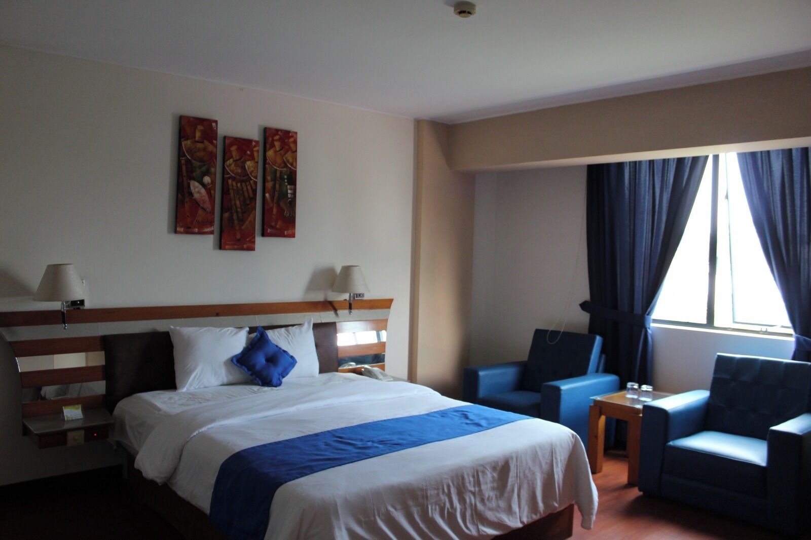 Habitación de hotel con una cama grande, dos sillones, una mesita y una ventana con cortinas.