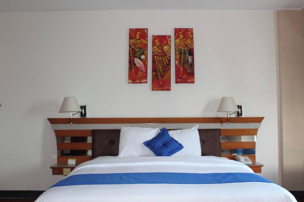 Camera d'albergo con un letto, due abat-jour, un poggiatesta in legno e tre quadri appesi al muro.