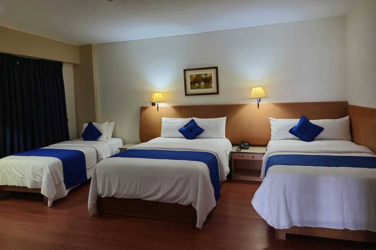 Camera d'albergo con tre letti, ognuno con biancheria bianca e dettagli blu, una testiera in legno, un comodino con una lampada e un quadro appeso al muro.