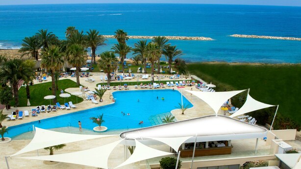 A resort with a large swimming pool, palm trees, and a view of the ocean. - Venus Beach Hotel (Paphos)