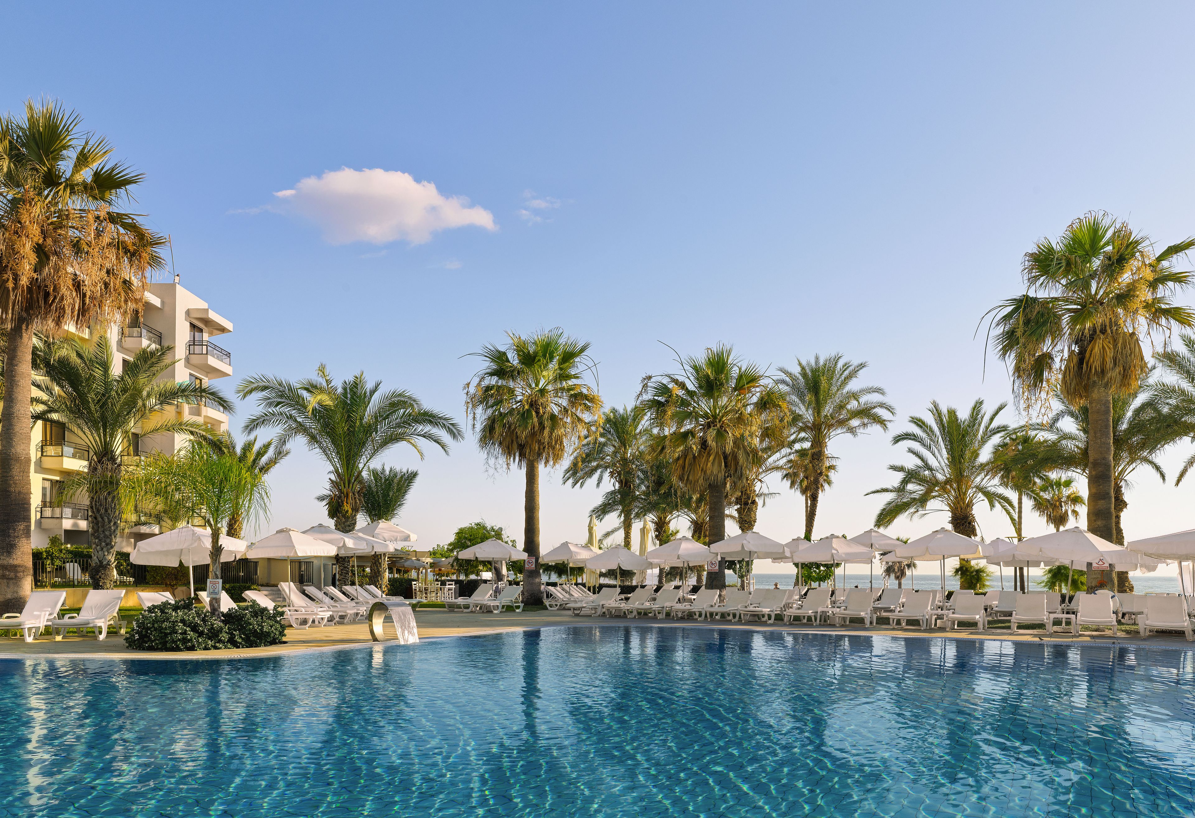 Zona de piscina con sillones de playa y sombrillas, rodeada de palmeras y un edificio.