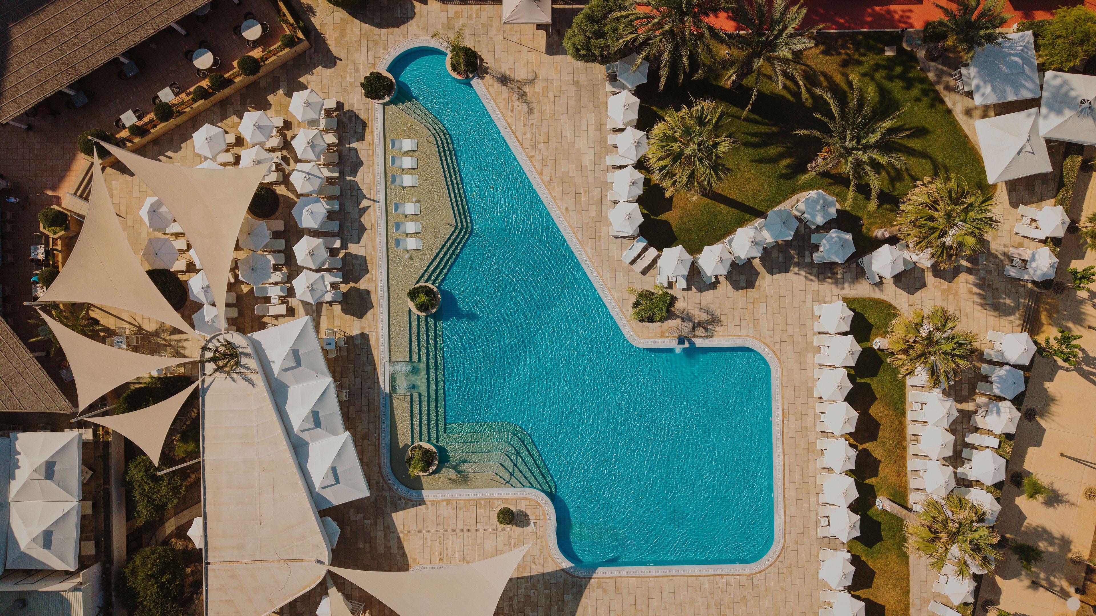 Eine Vogelperspektive auf einen Swimmingpool, umgeben von Liegestühlen und Sonnenschirmen, mit blauem Himmel darüber.