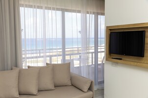 Una habitación con sofá color beige, televisor montado sobre un marco de madera, y un ventanal con vista a la playa y al océano.