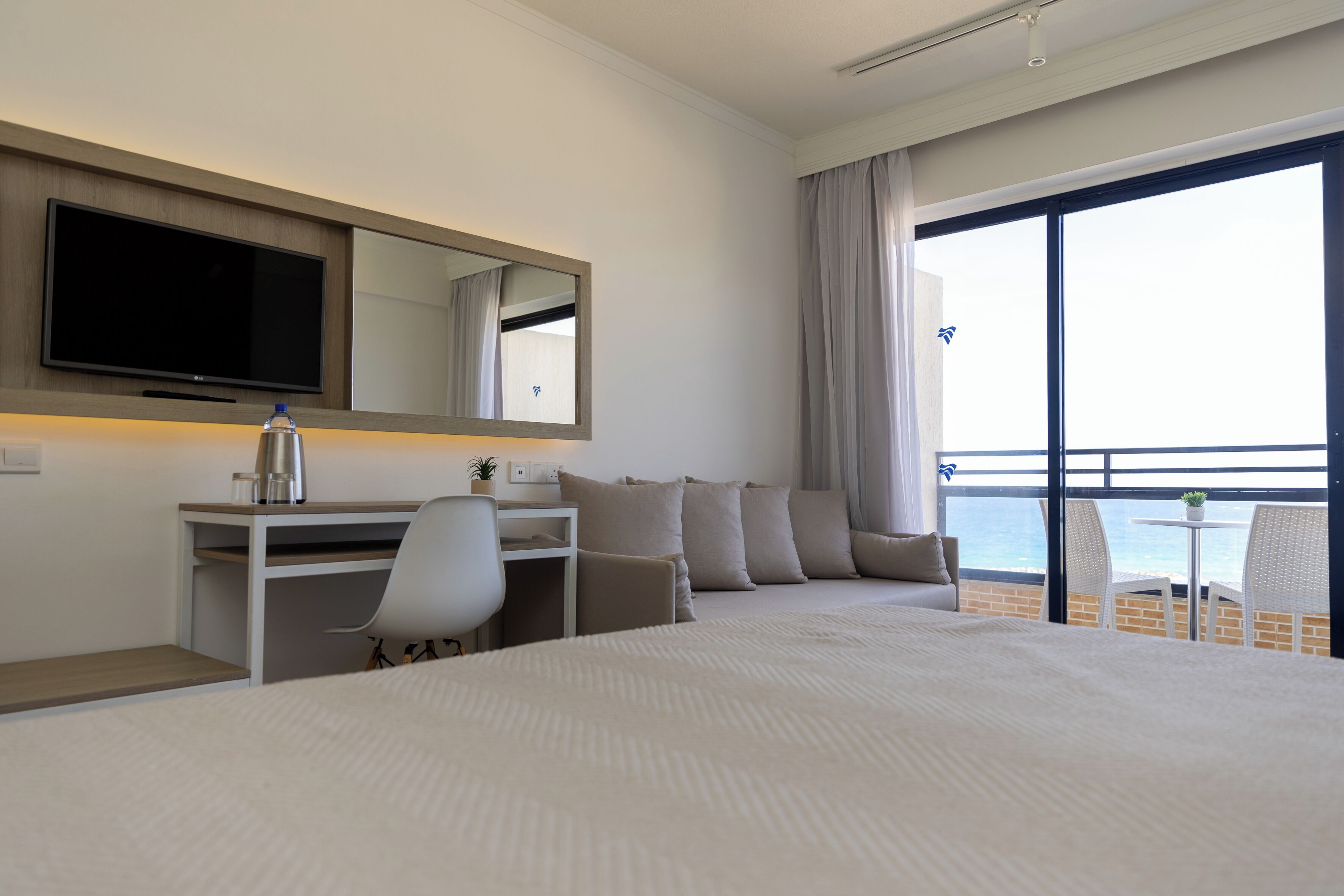 A hotel room with a large bed, a desk with a chair, a TV, and a balcony with a view of the sea.