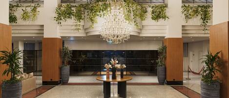 A lobby with a chandelier, potted plants, a reception desk, and a glass door entrance.
