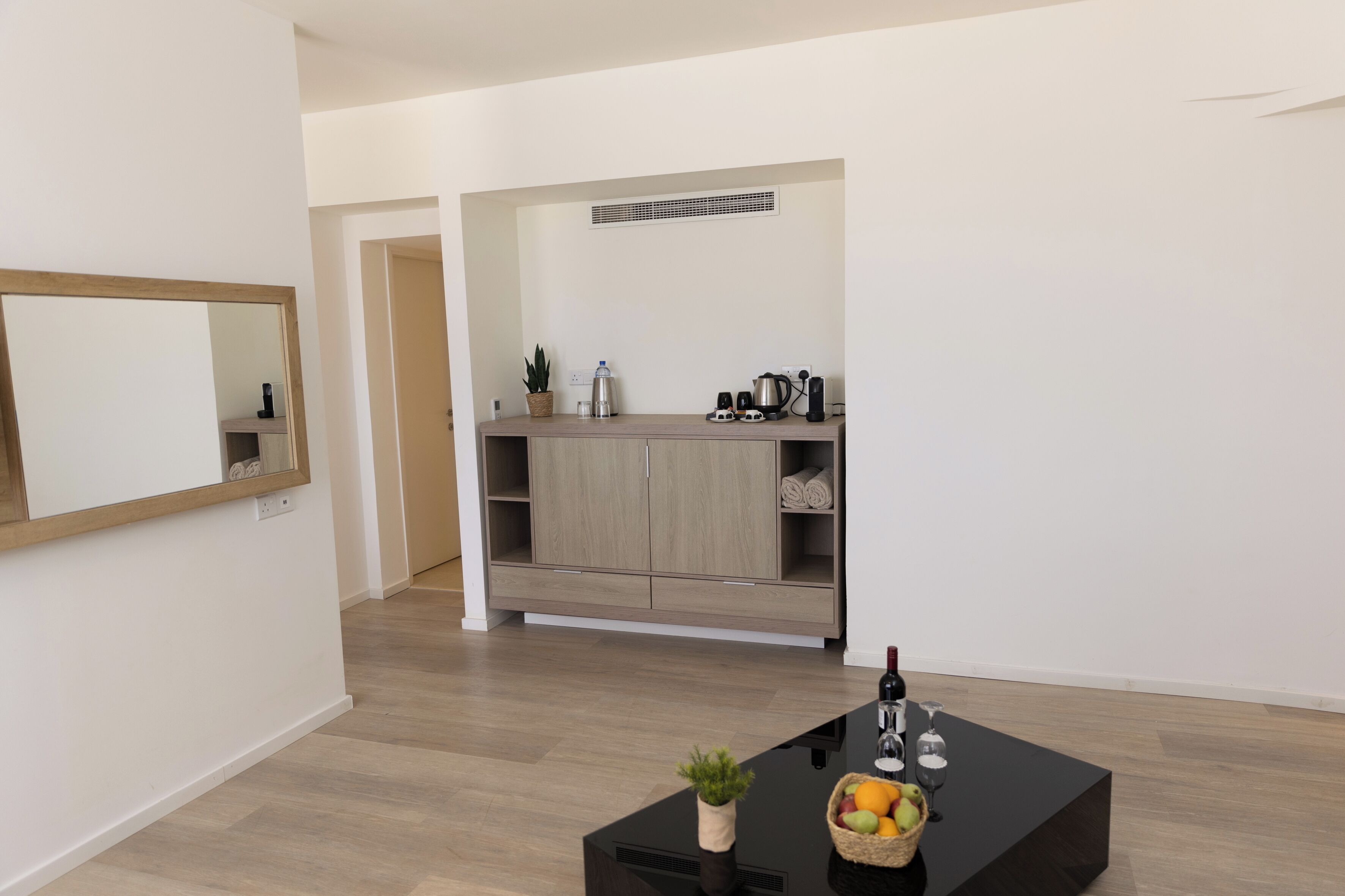 A modern living room with a black coffee table, a small plant, and a fruit basket. There is a kitchen area with wooden cabinets and a sink.