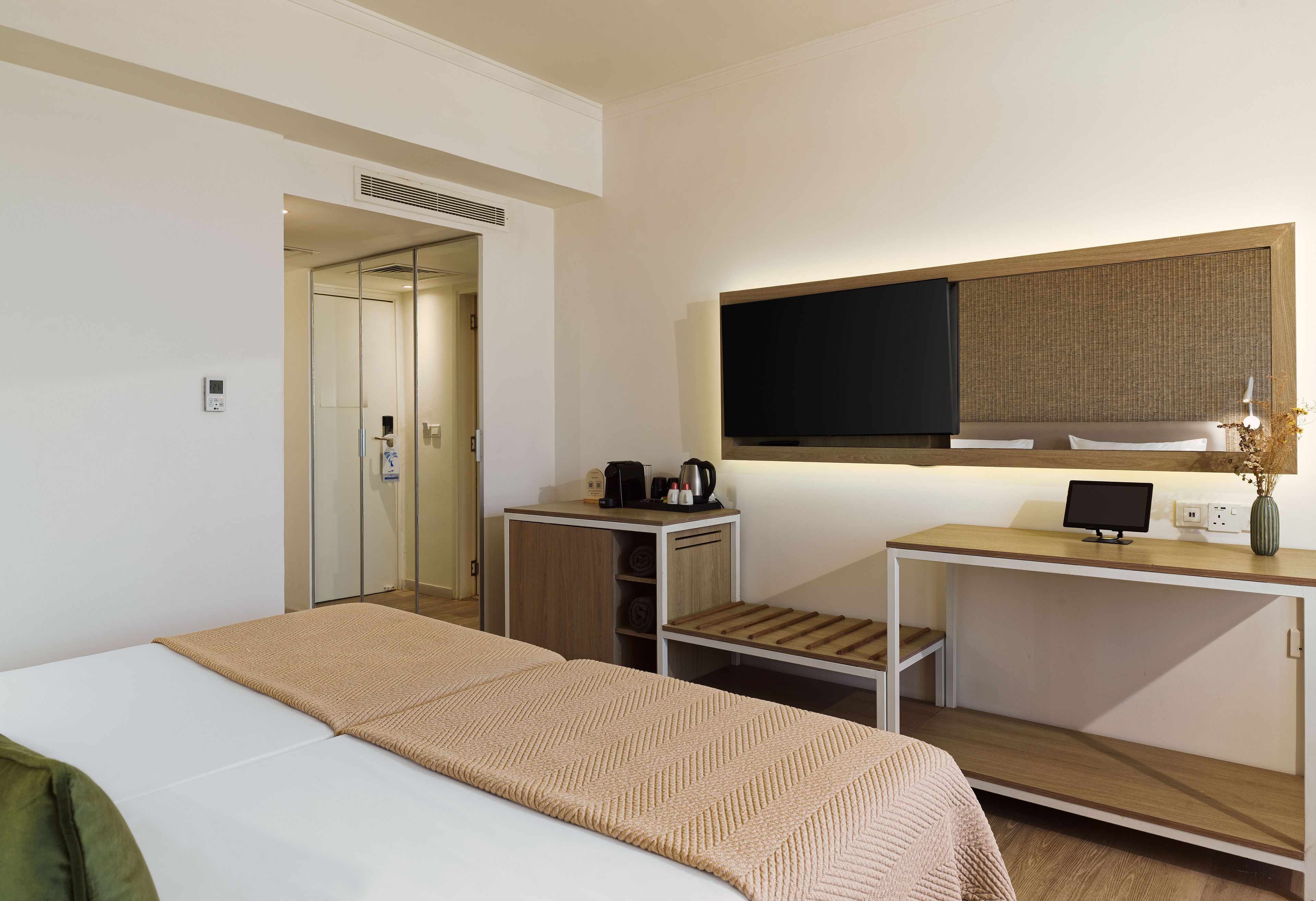 A hotel room with a bed, a desk with a flat-screen TV, a coffee maker, and a small shelf.