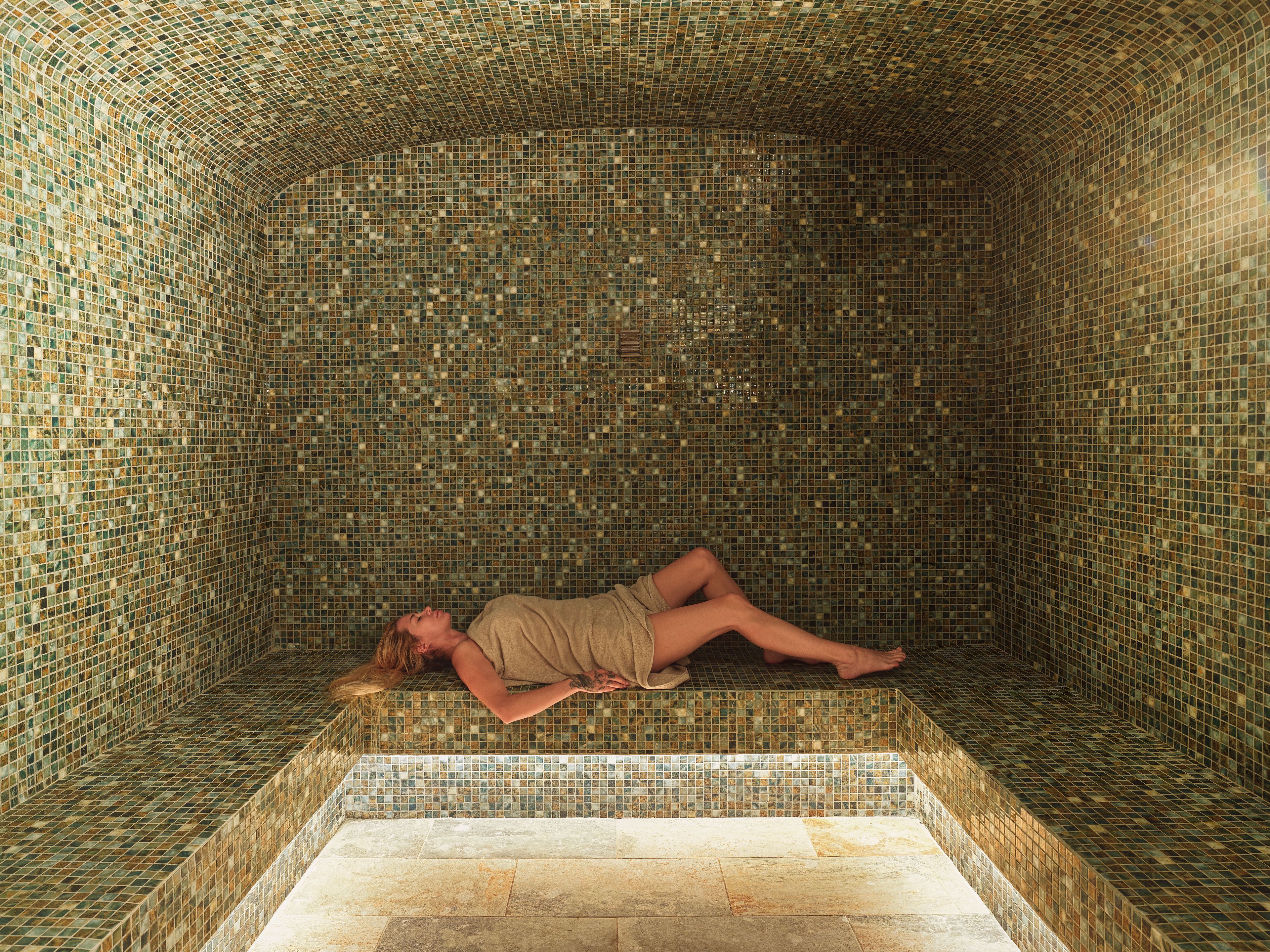 A room with a tiled floor and walls, a bench, and a person lying on the bench.