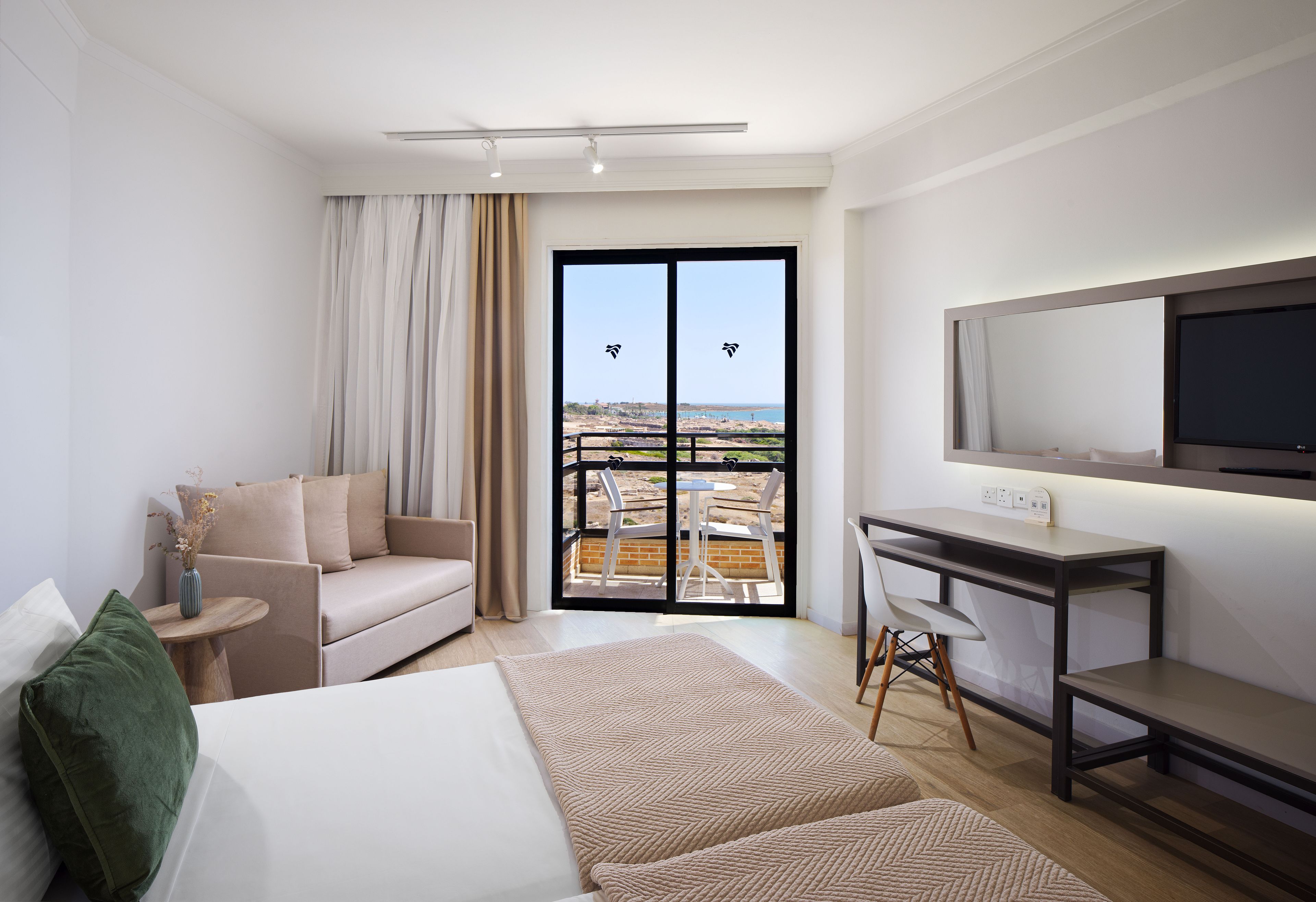 A hotel room with a bed, a desk, a chair, a sofa, a balcony with a view, and a television.