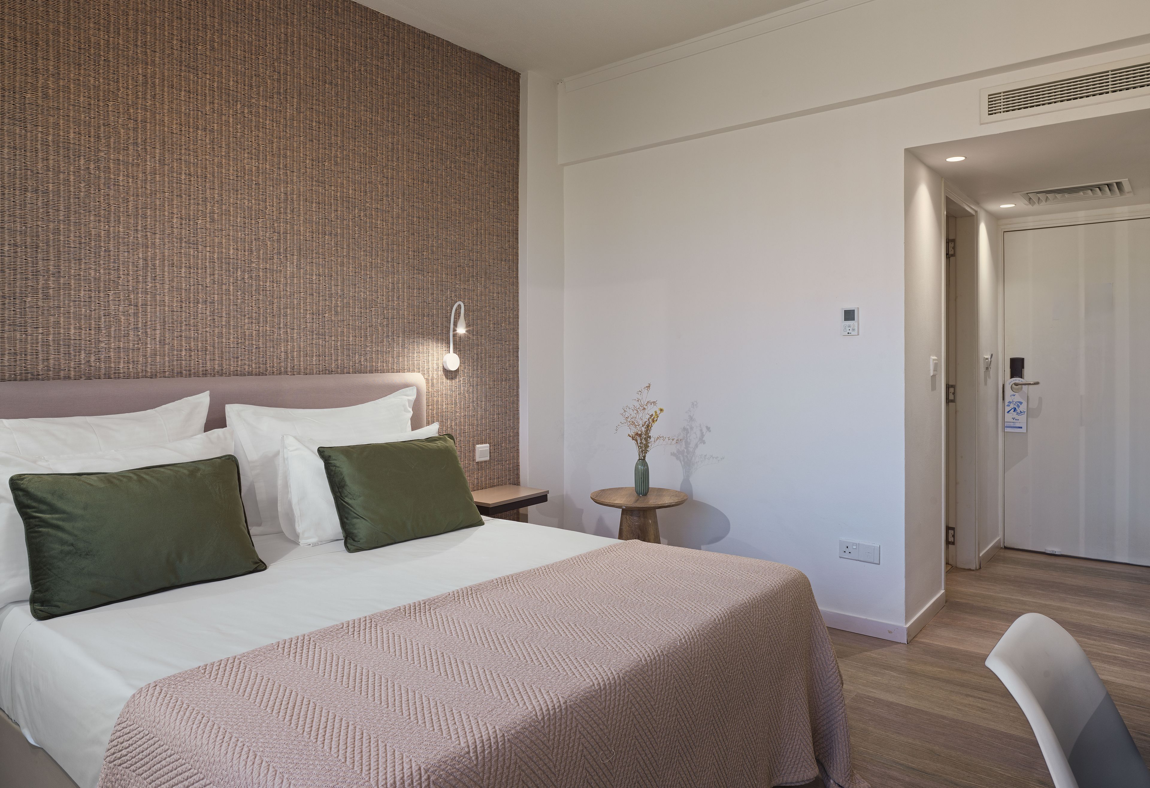 A hotel room with a bed, pillows, a bedside table with a vase, and a door leading to another room.