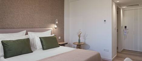 A hotel room with a bed, pillows, a bedside table with a vase, and a door leading to another room.