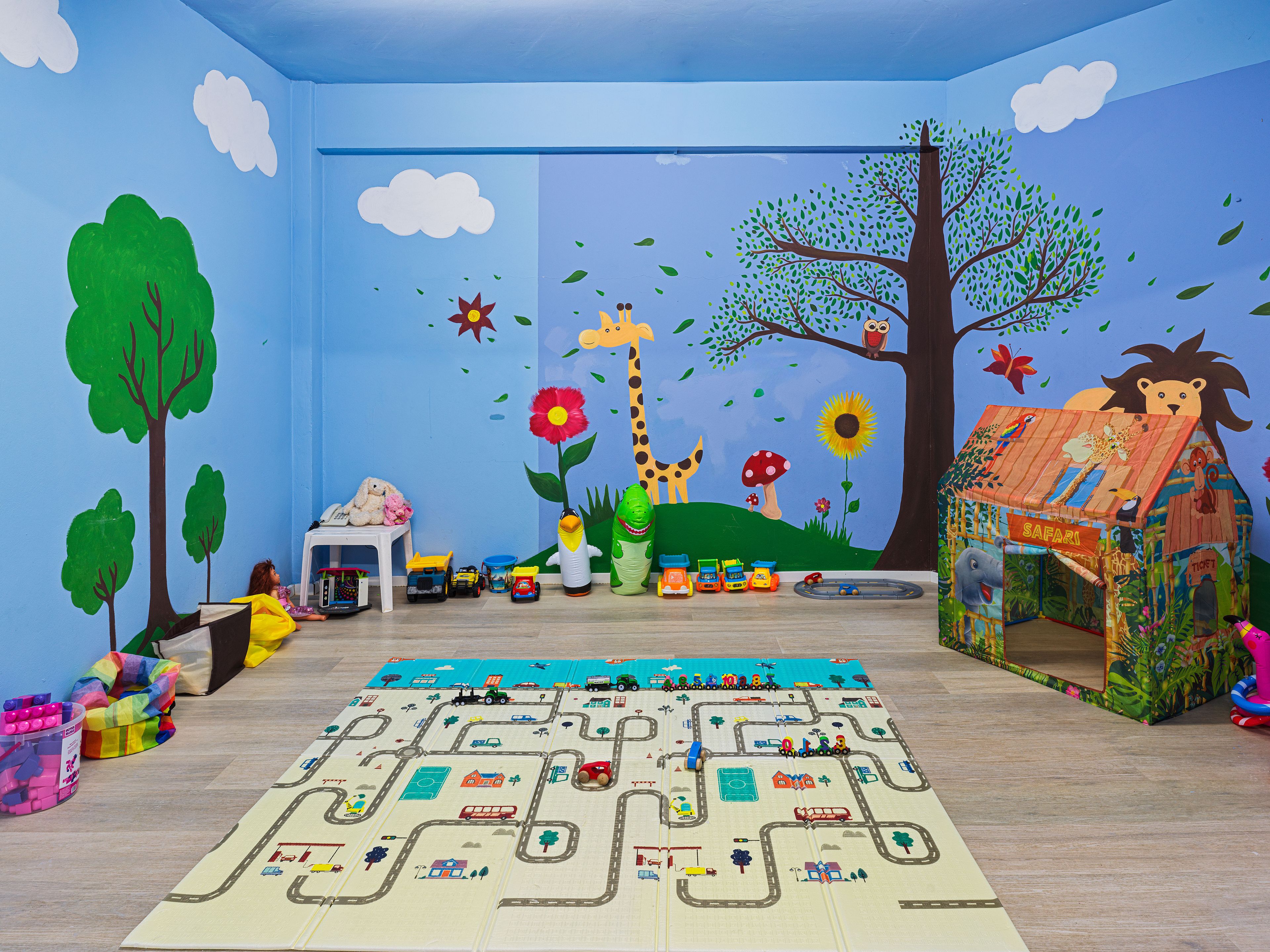 Un cuarto de juegos infantil con un tapete de juego grande con temática de safari, juguetes y un mural con animales y un árbol.