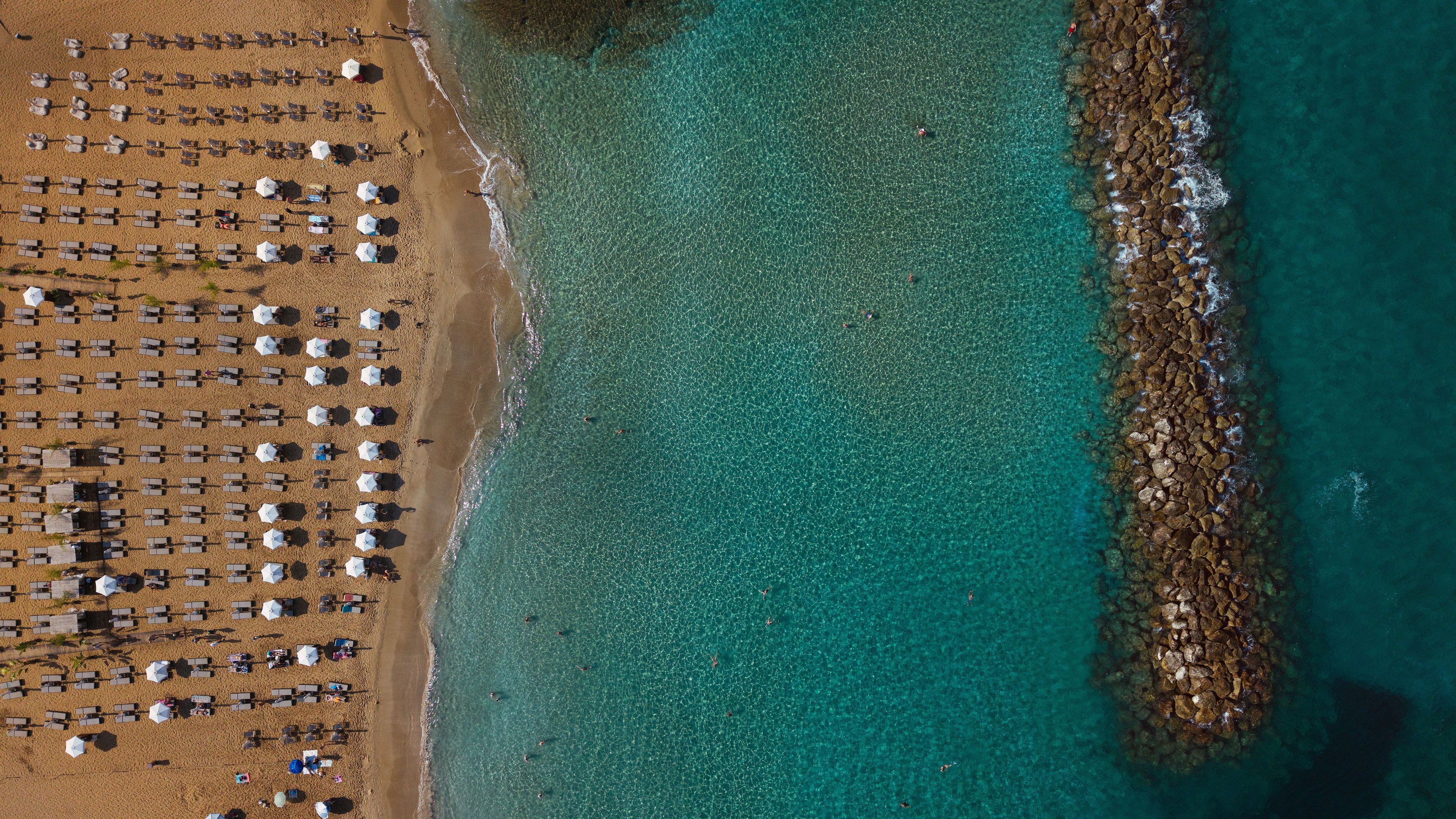 An aerial view of a beach with rows of sunbeds and umbrellas, clear turquoise water, and a rocky shoreline.