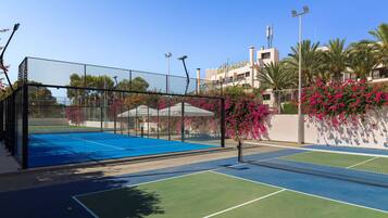 Cancha de tenis exterior con red, rodeada por una cerca y adyacente a un edificio con balcón.